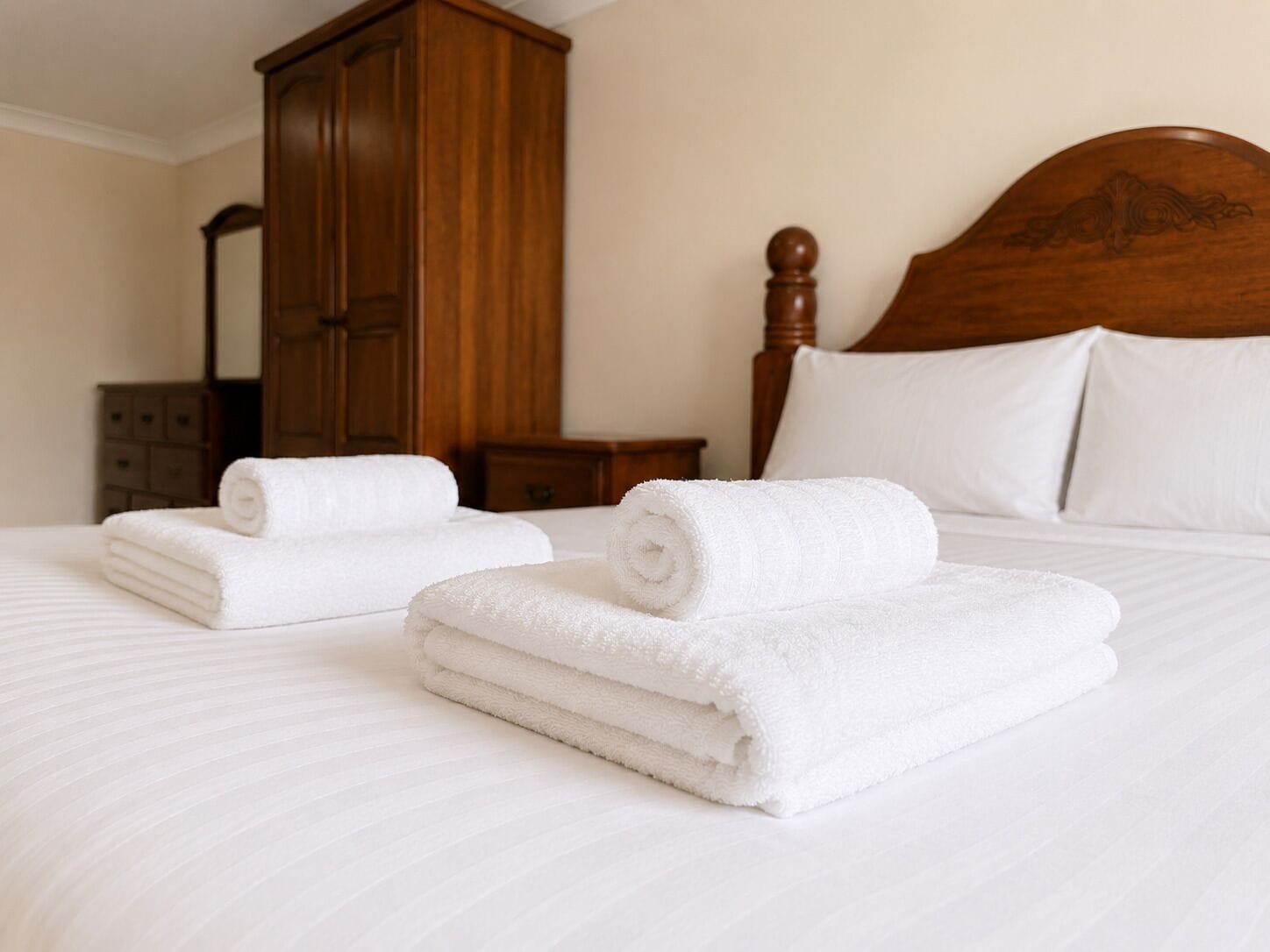 A neatly made bed with white linens and two rolled towels, a wooden wardrobe, and a nightstand with a lamp.