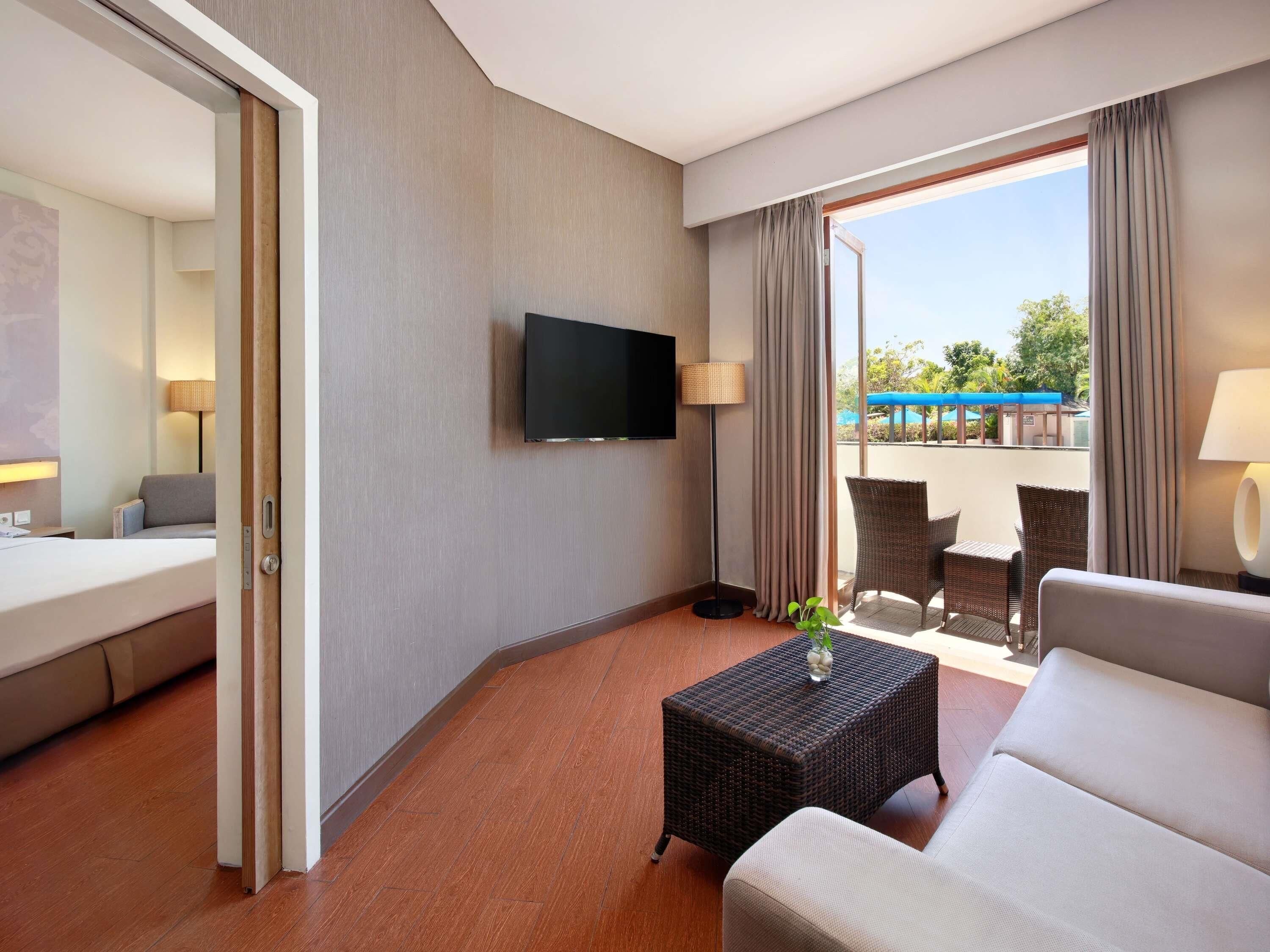 A hotel room with a bed, a television, a small table with a plant, a sofa, and a balcony with outdoor furniture.