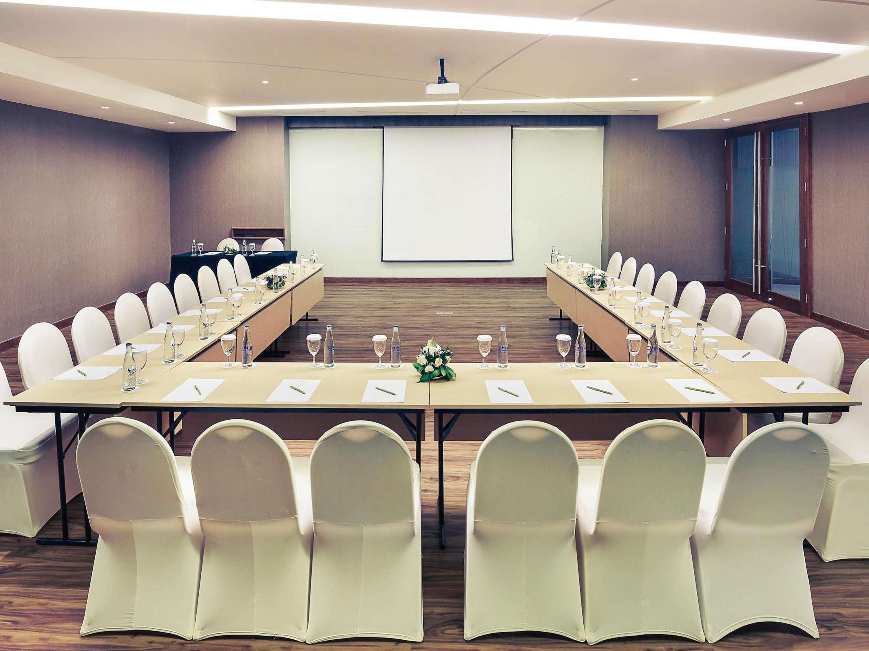 A conference room with a long table, white chairs, water bottles, and a projector screen.