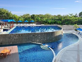 Una gran piscina curvo con sillas de descanso y sombrillas, rodeada de follaje y un área de terraza con baldosas.