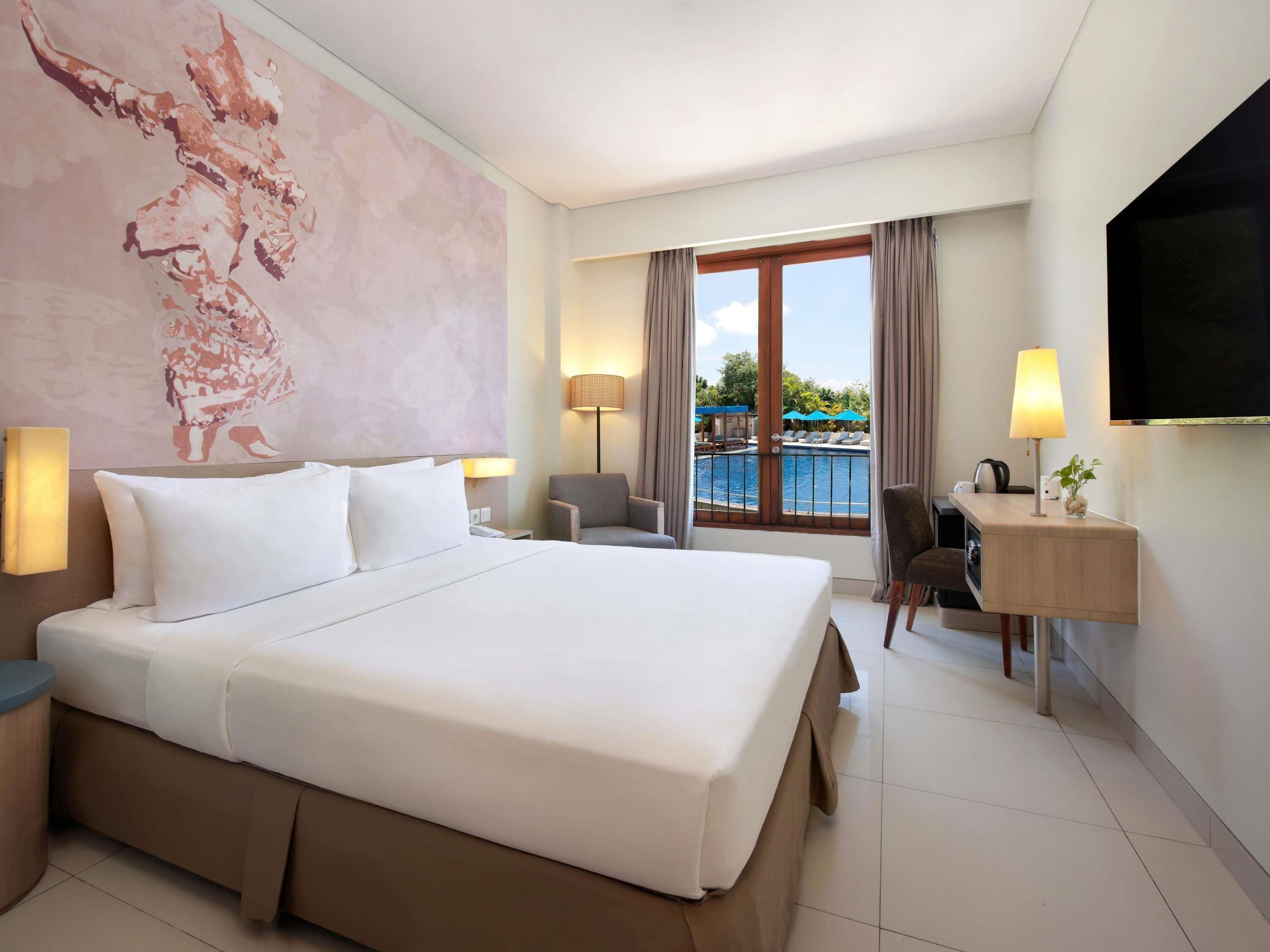 Ein Hotelzimmer mit einem großen Bett, einem Schreibtisch, einem Sessel, einem Fernseher, einer Lampe und Blick auf einen Pool.