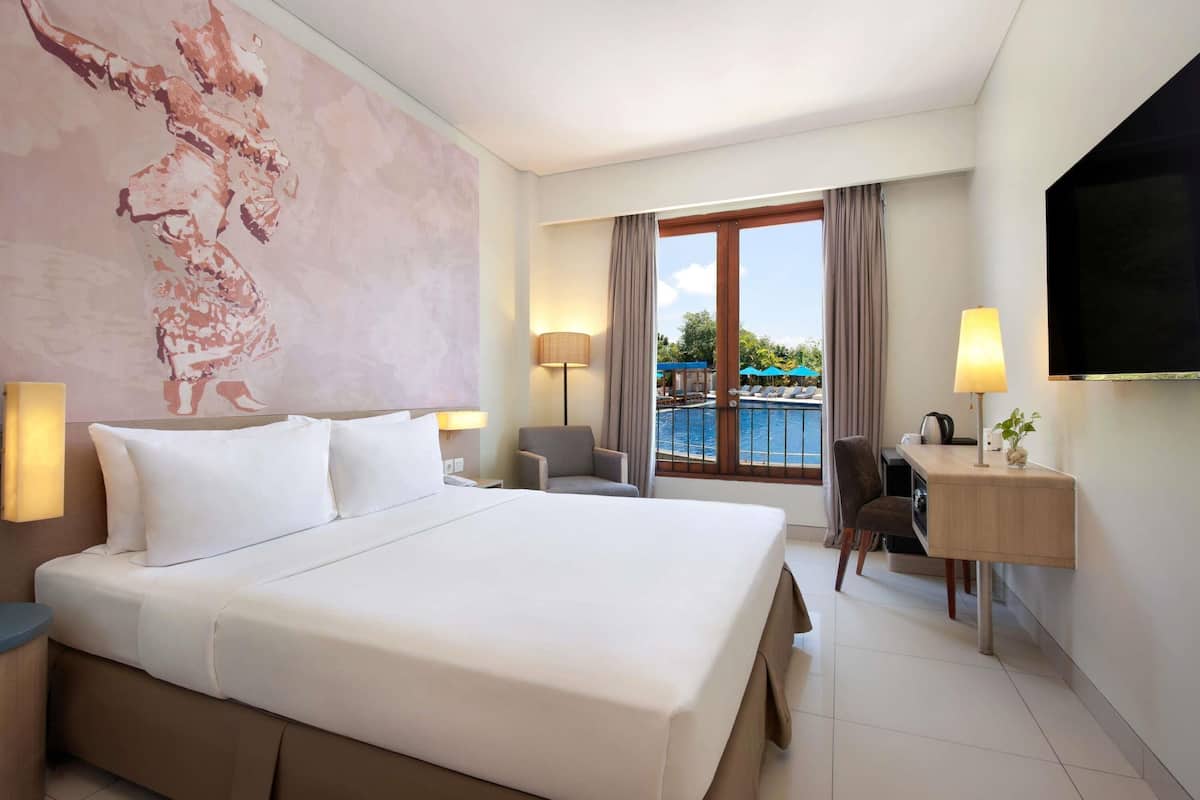 Ein Hotelzimmer mit einem großen Bett, einem Schreibtisch, einem Sessel, einem Fernseher, einer Lampe und Blick auf einen Pool.