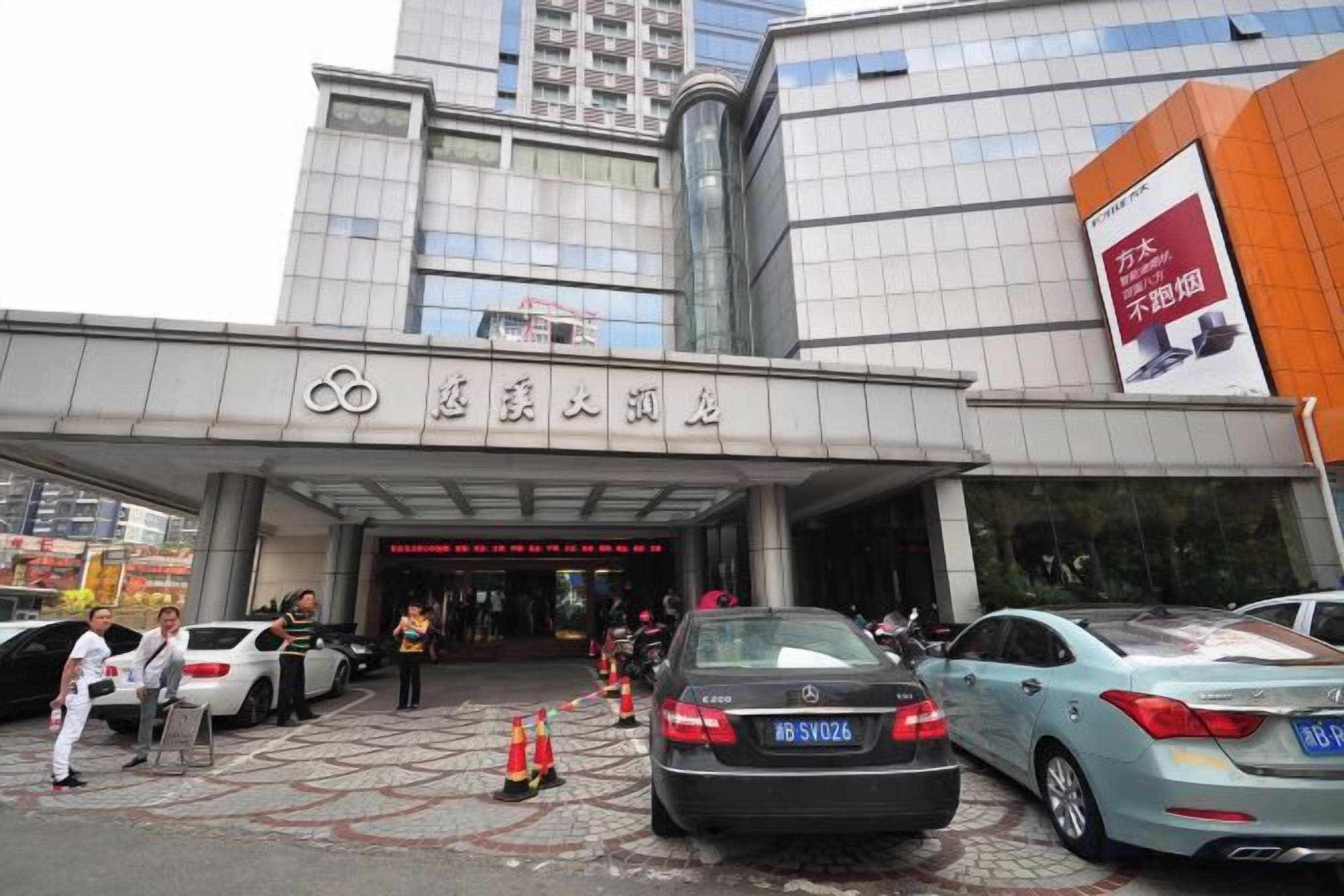 A building entrance with a sign, cars parked in front, and a few people.