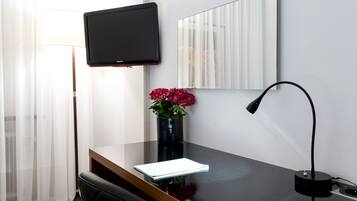 Una habitación de hotel moderna con una televisión de pantalla plana montada en la pared, un escritorio con una computadora portátil, un florero con flores, una lámpara y una silla.