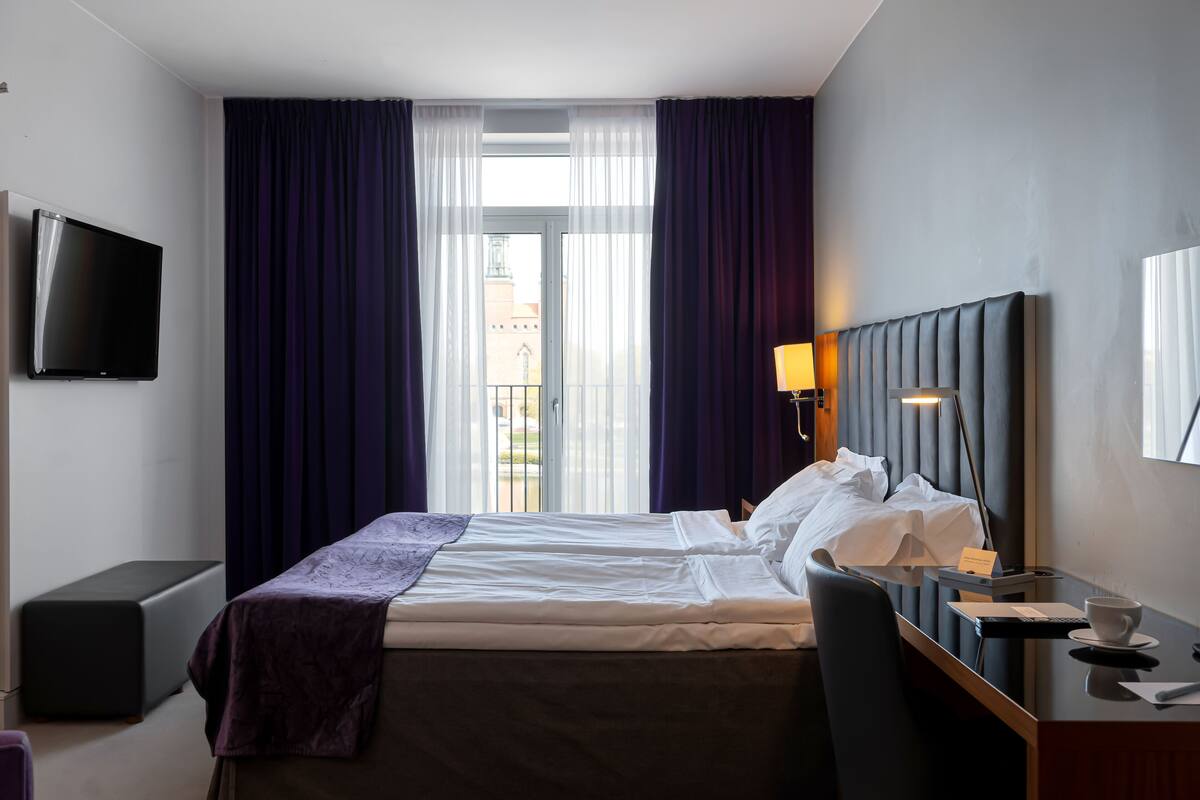 Ein Hotelzimmer mit Bett, Schreibtisch, Stuhl, Fernseher und einem Fenster mit Vorhängen.