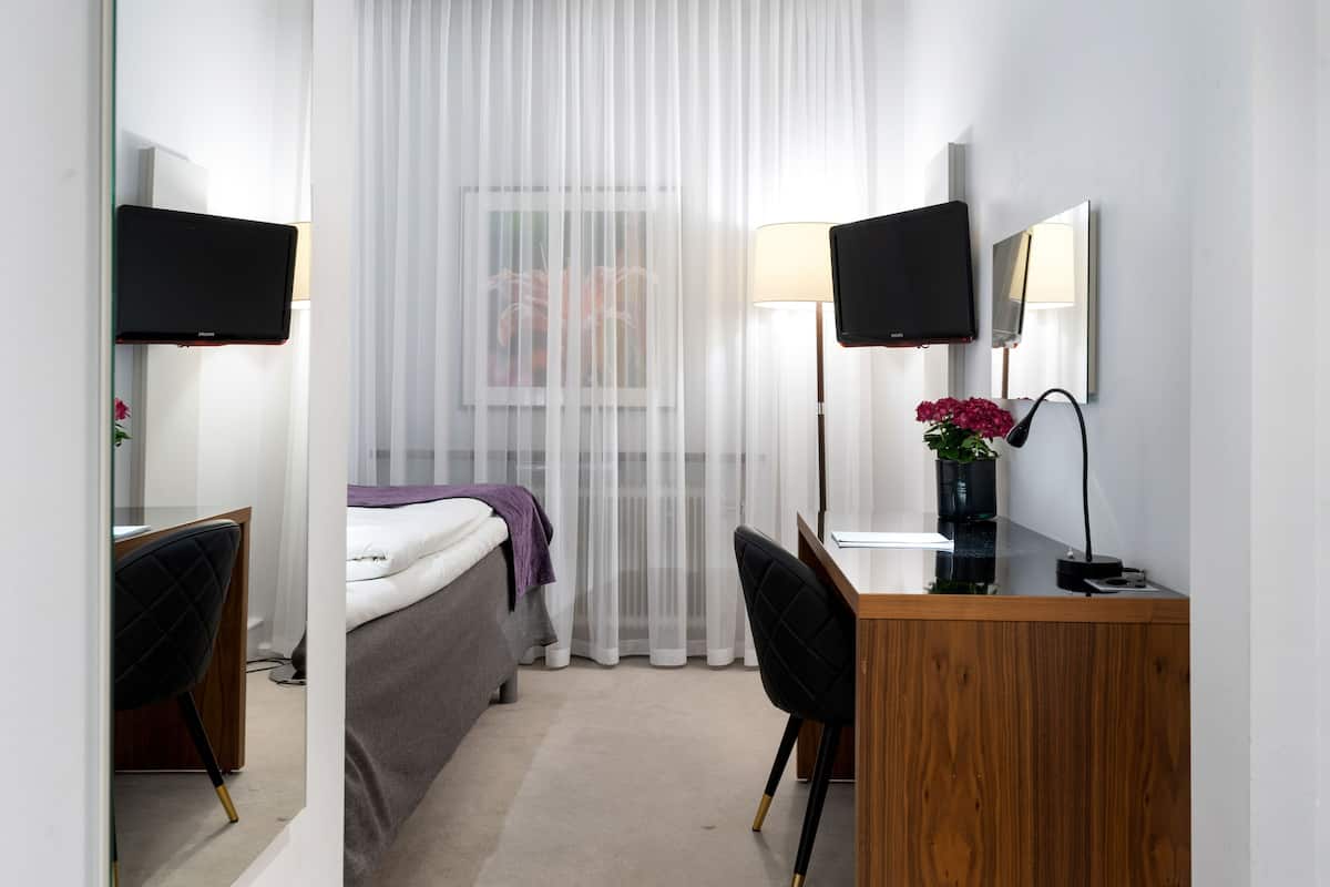 Ein Hotelzimmer mit einem Bett, einem Schreibtisch mit Computer, zwei Monitoren, einer Lampe und einer Vase mit Blumen.