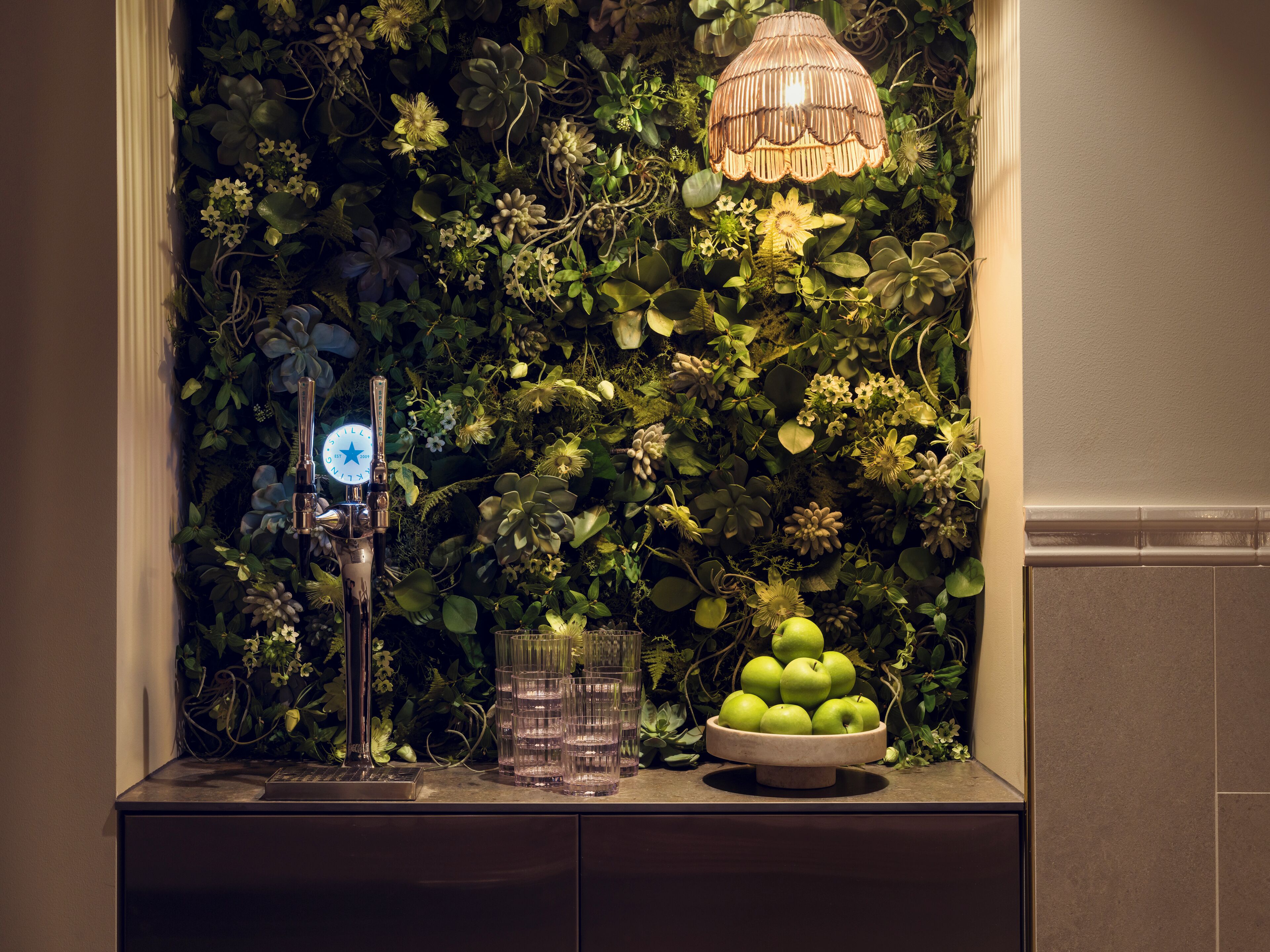 A bar area with a decorative wall, a bar tap, glasses, and a bowl of apples.
