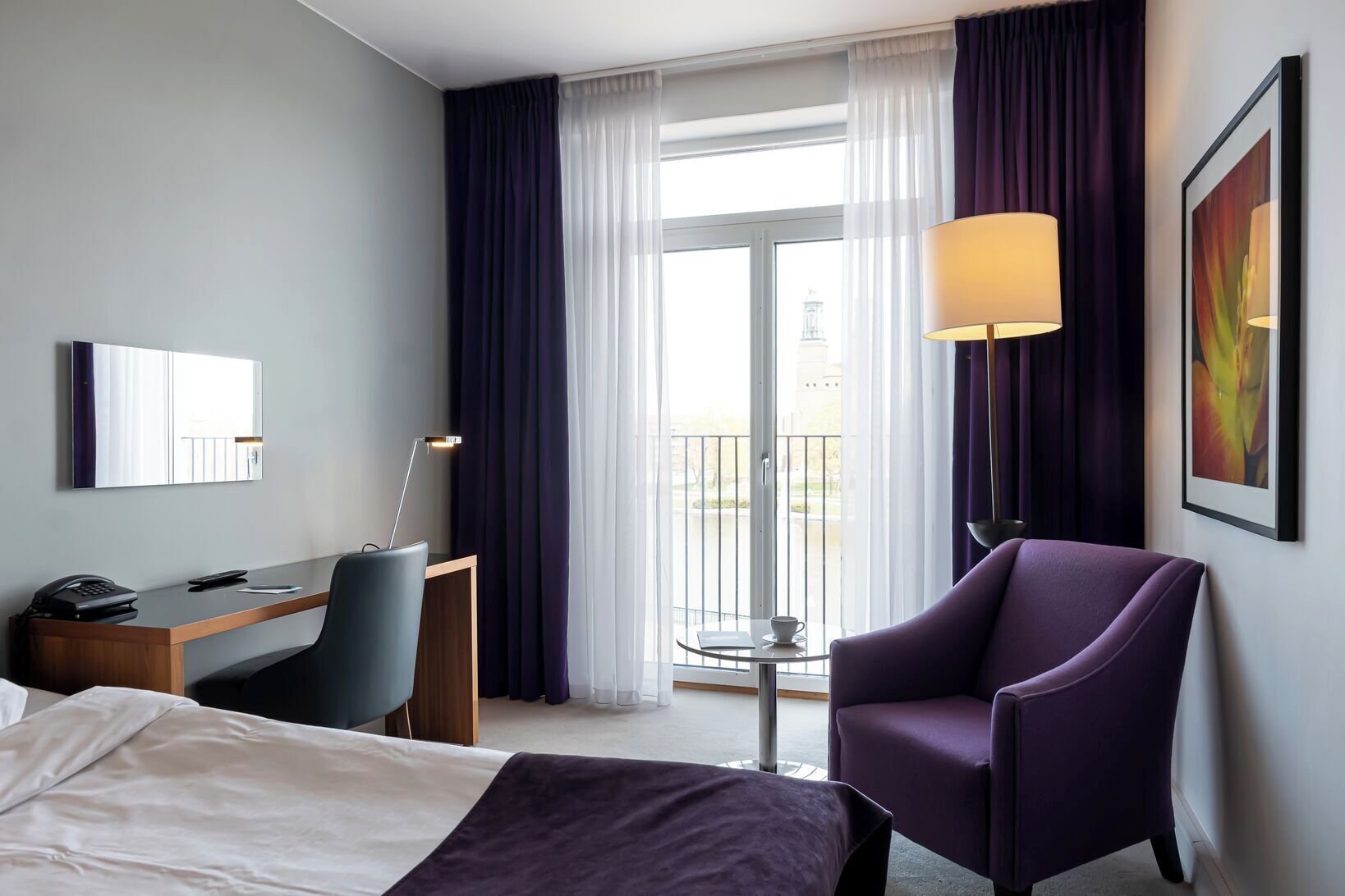 A hotel room with a bed, a desk with a chair, a purple armchair, a lamp, and a window with curtains.