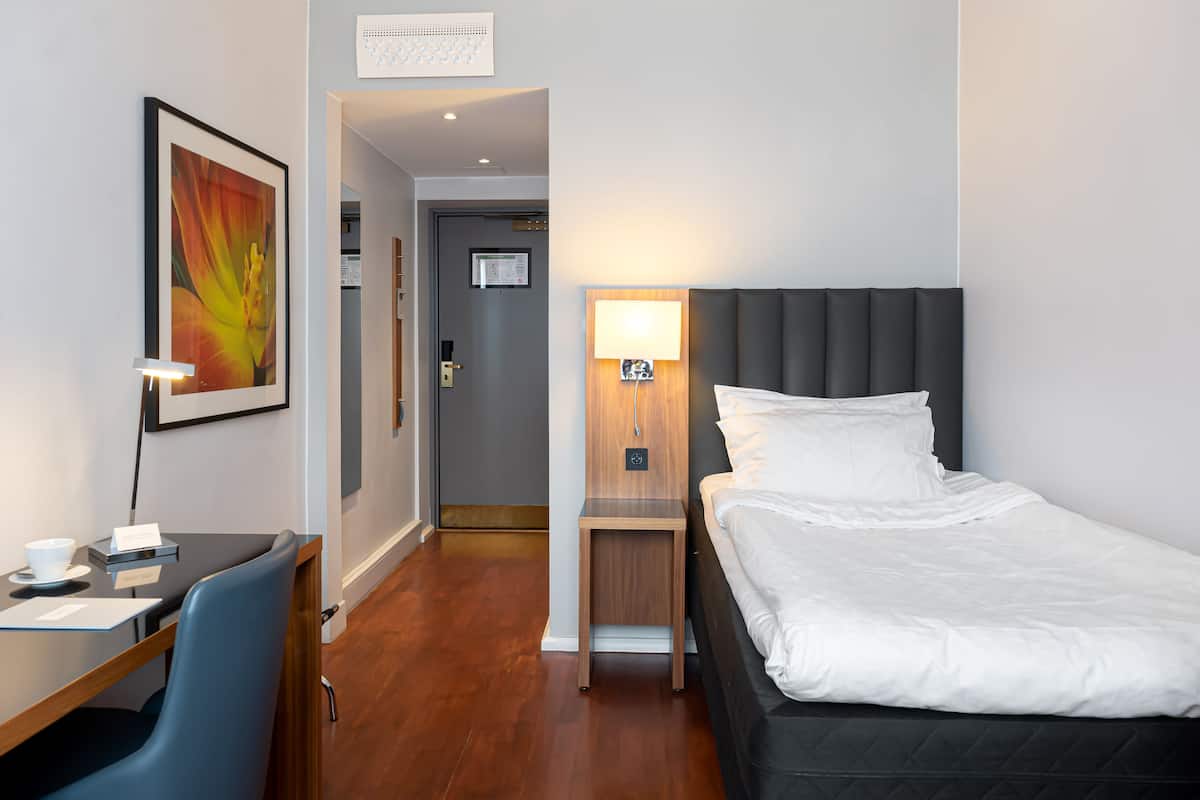 Ein Hotelzimmer mit Bett, Schreibtisch mit Stuhl, einem kleinen Tisch mit Lampe und einem gerahmten Bild an der Wand.