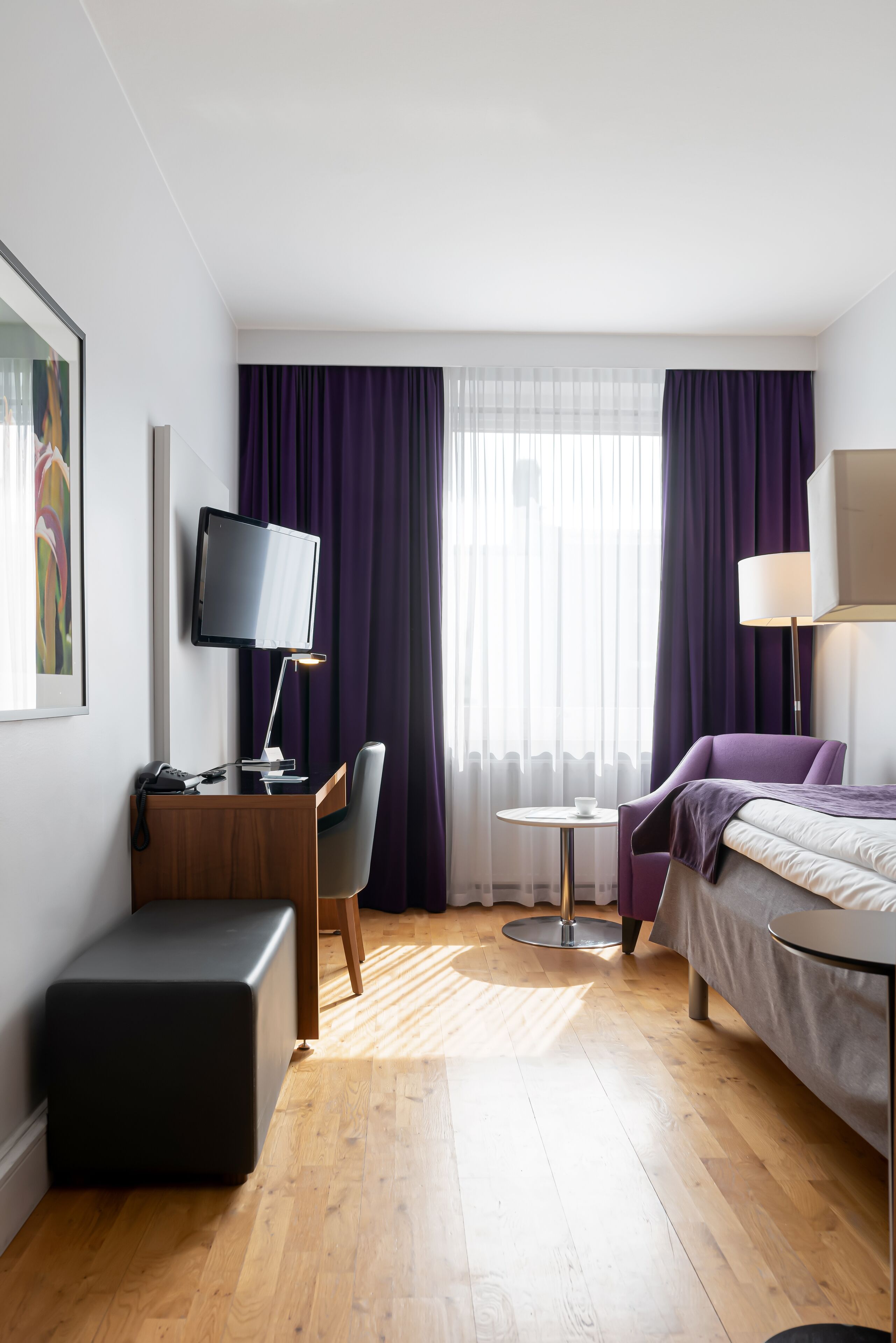 A hotel room with a bed, a desk with a computer, a chair, a small table, and a lamp.
