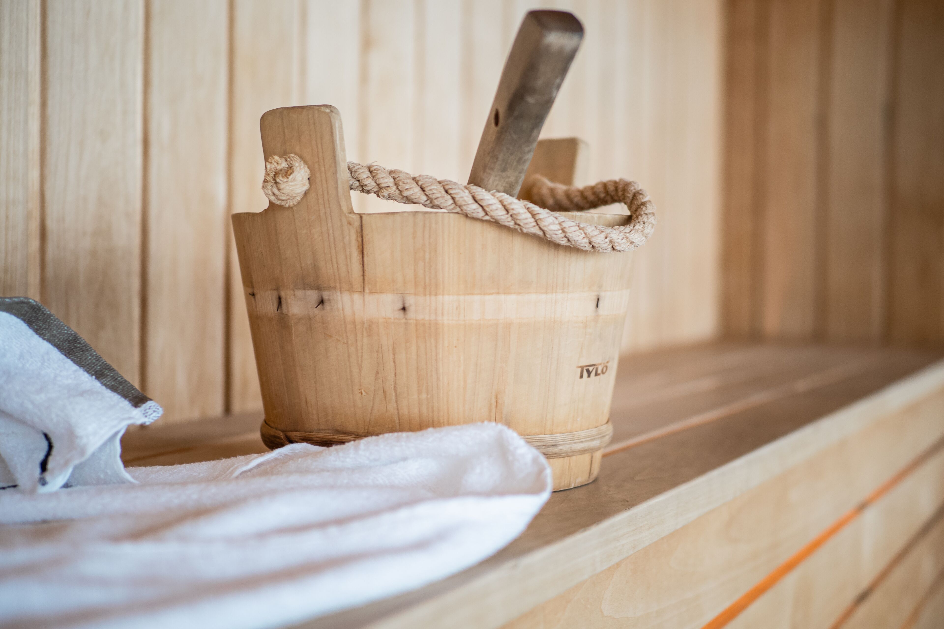 A wooden sauna with a rope handle and a metal tool inside, placed on a wooden shelf with a white towel beside it.
