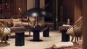 Un espace de détente moderne avec un foyer central, des meubles en osier et un pouf à carreaux.