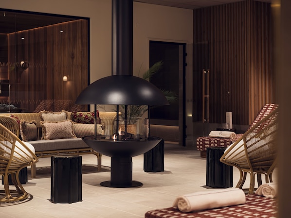 Un espace de détente moderne avec un foyer central, des meubles en osier et un pouf à carreaux.