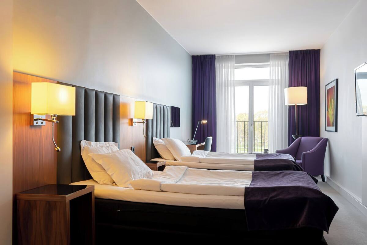 Ein Hotelzimmer mit zwei Betten, einem Schreibtisch, einem Stuhl und einem Balkon mit Vorhängen.