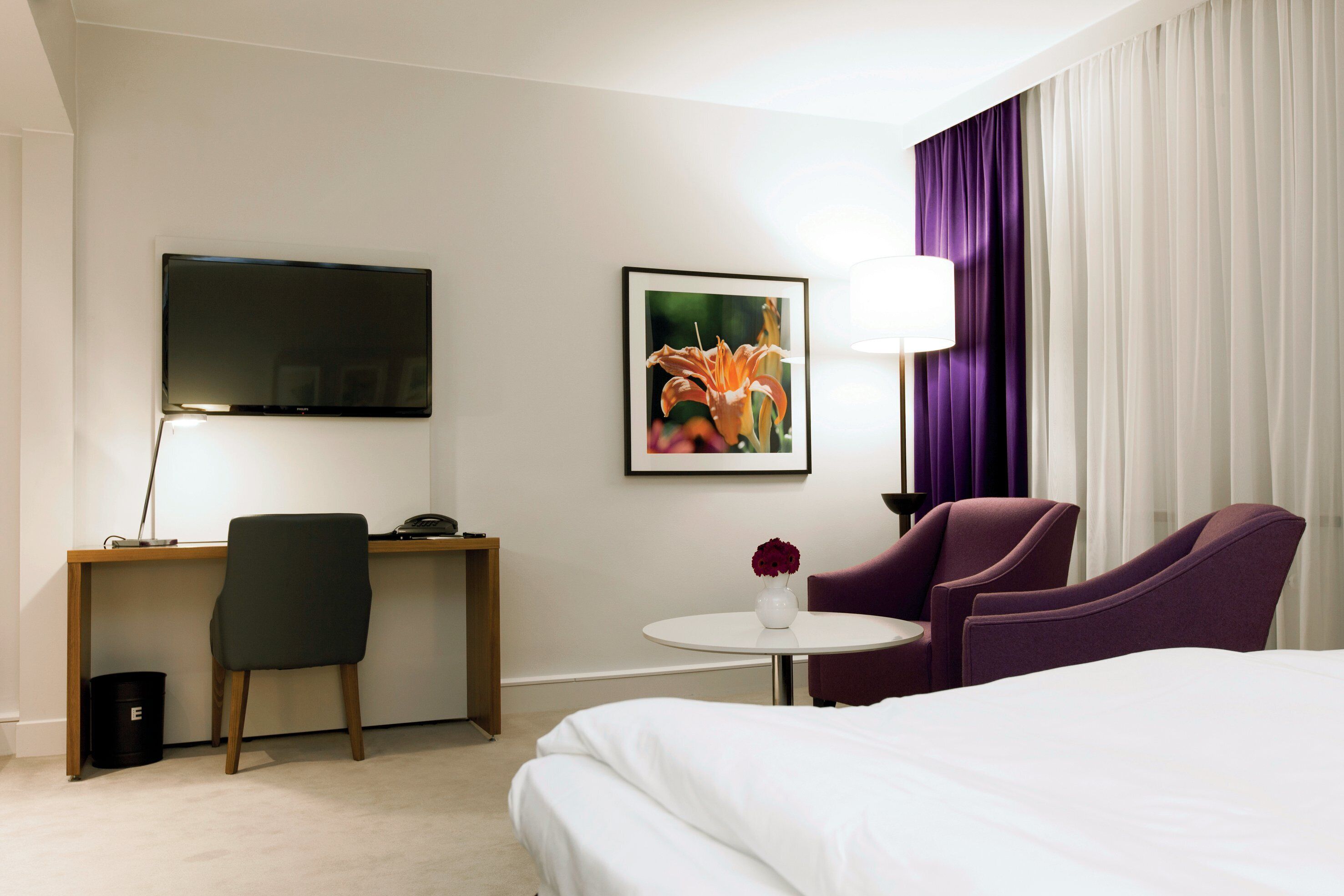 A hotel room with a bed, a desk with a chair, a television, a framed picture, a small table with a vase, and purple armchairs.