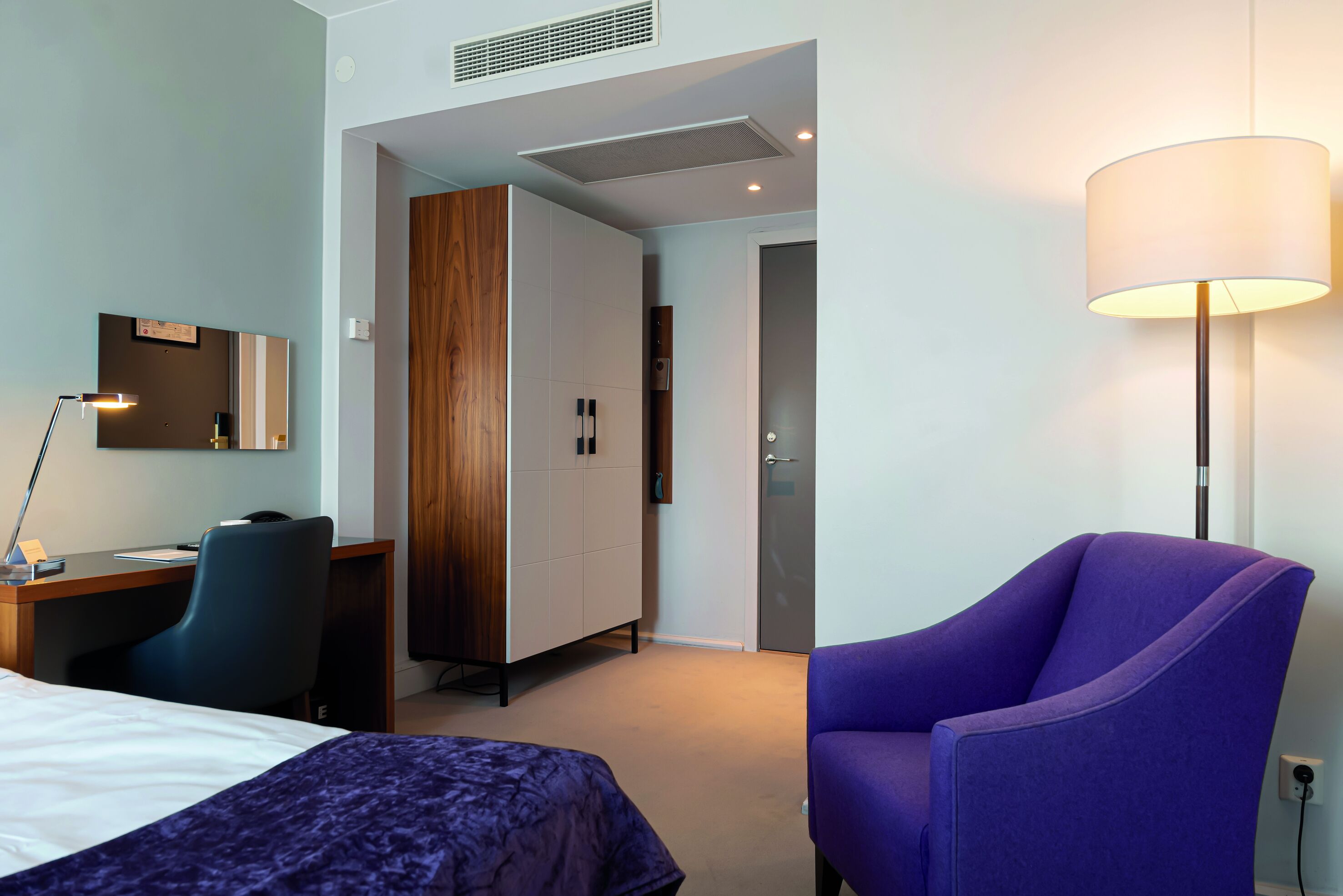 Una habitación de hotel con cama, escritorio, silla, armario y lámpara.