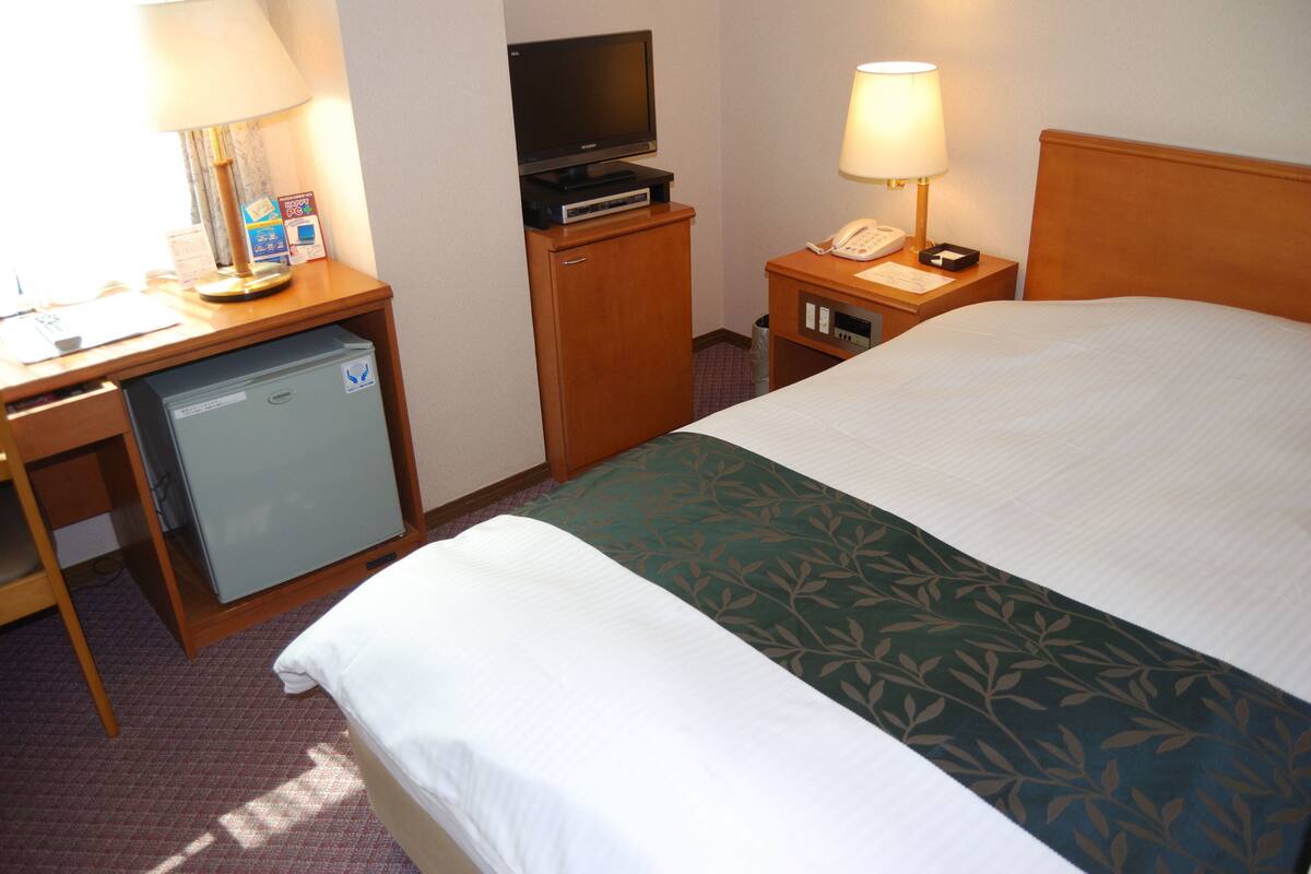 Ein Hotelzimmer mit einem Bett, einem Schreibtisch mit Fernseher, einem Minikühlschrank, einer Lampe und einem Telefon.