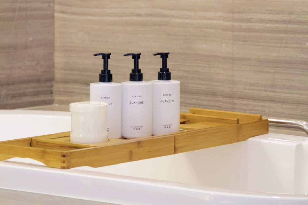 A bathroom with a wooden tray holding three white bottles of BYREDO BLANCHE hair care products and a white candle.