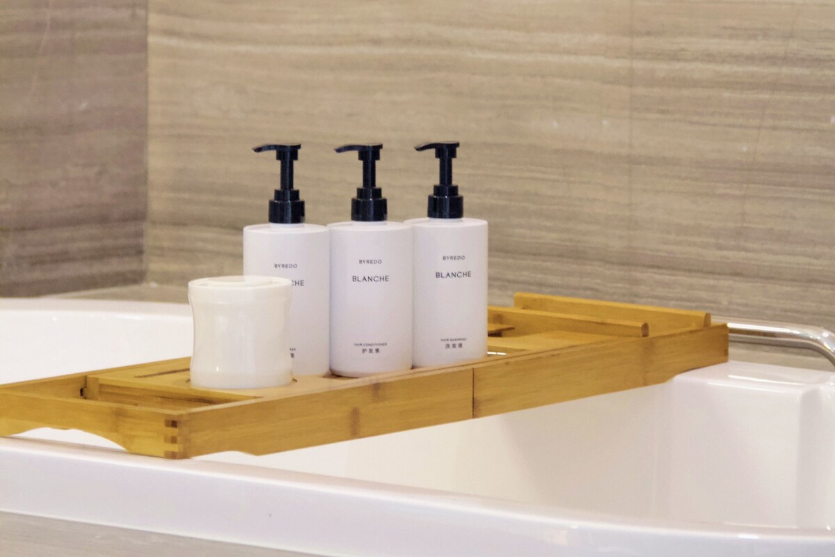 A bathroom with a wooden tray holding three white bottles of BYREDO BLANCHE hair care products and a white candle.