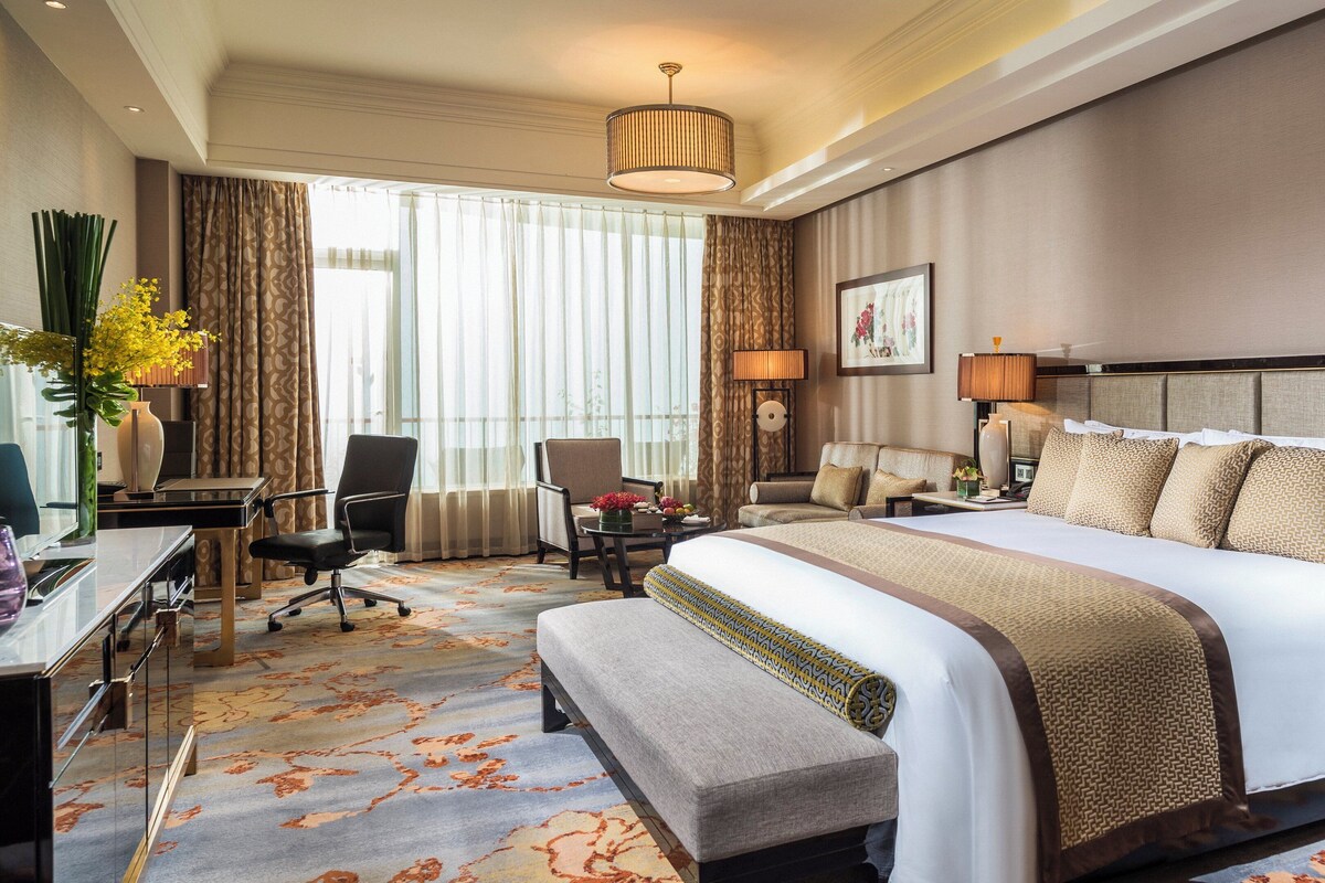 A hotel room with a large bed, a desk with a chair, a sofa, a bench, and a large window with curtains.