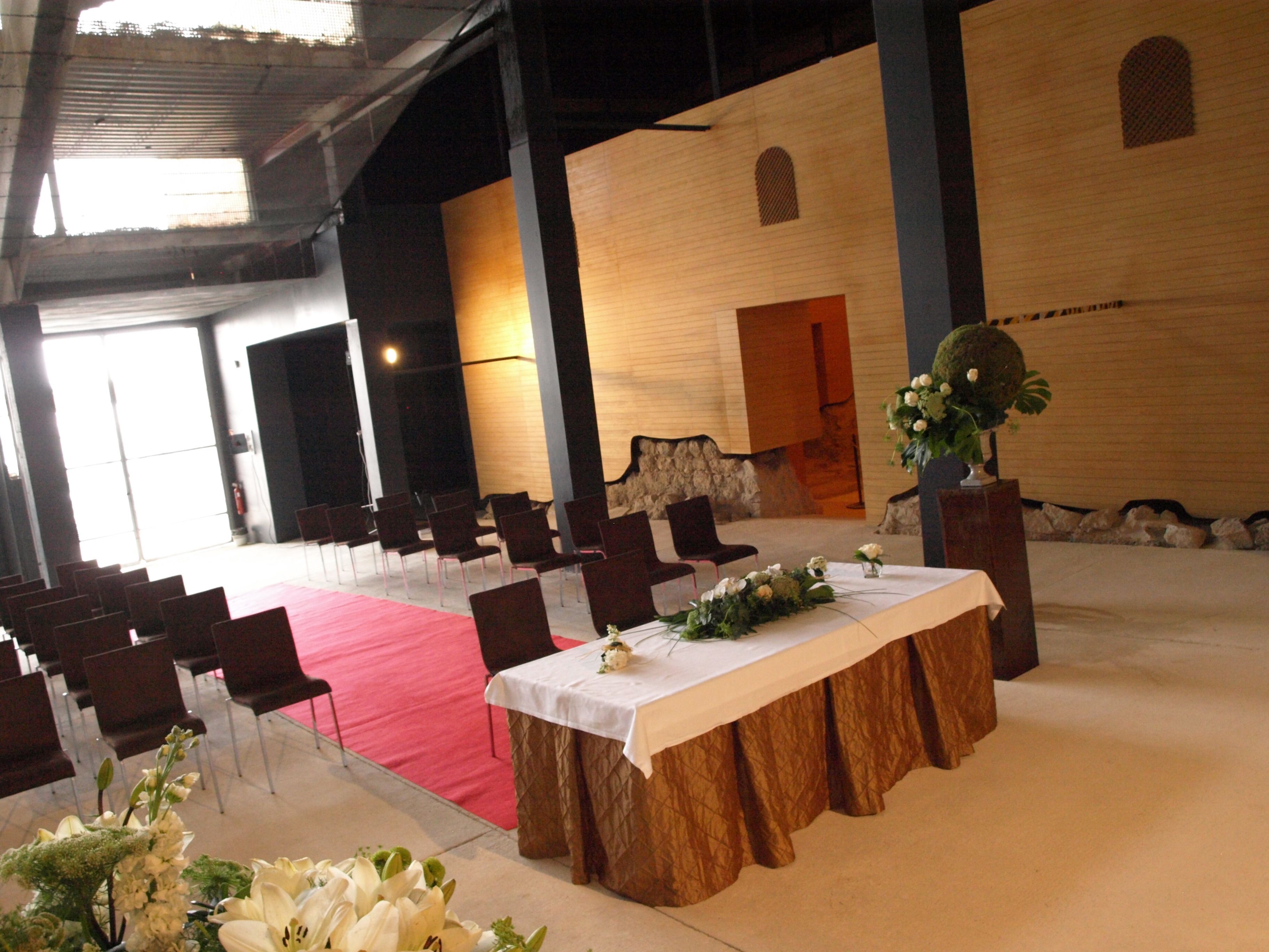 Un amplio salón con filas de sillas, una mesa central con mantel blanco y una alfombra roja.