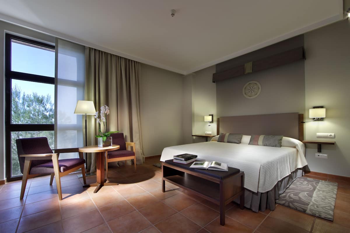 Habitación de hotel con cama, dos sillas, una mesita y ventana con cortinas.