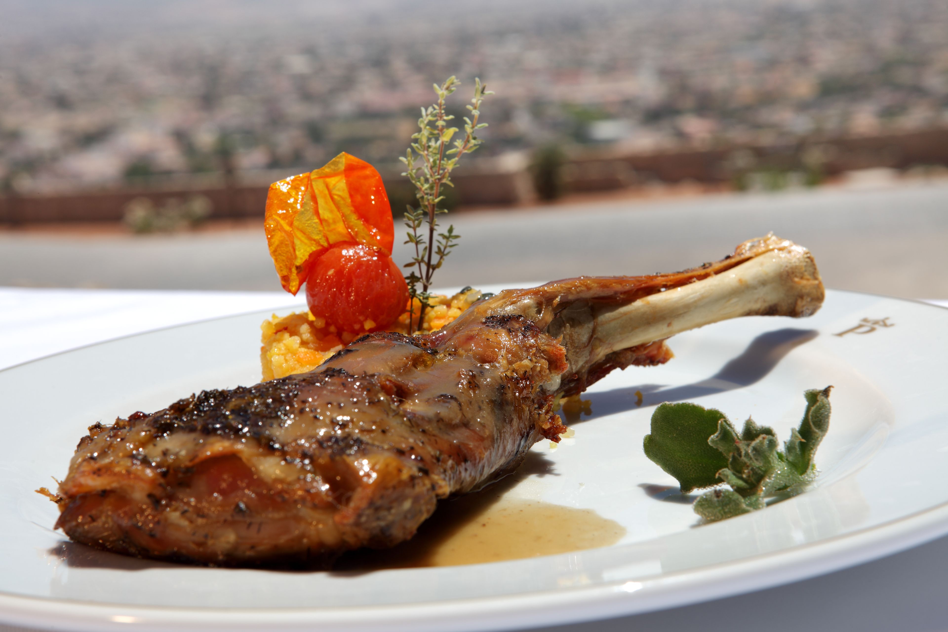 A plate with a grilled lamb chop, a roasted tomato, and a sprig of thyme, set against a cityscape background.
