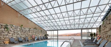 Una piscina interior con techo de cristal, paredes de piedra y sillones de descanso.