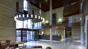 A spacious lobby with a high ceiling, a central seating area with a brown leather sofa, a reception desk, and a staircase leading to an upper level. - Parador de Lorca (Lorca)