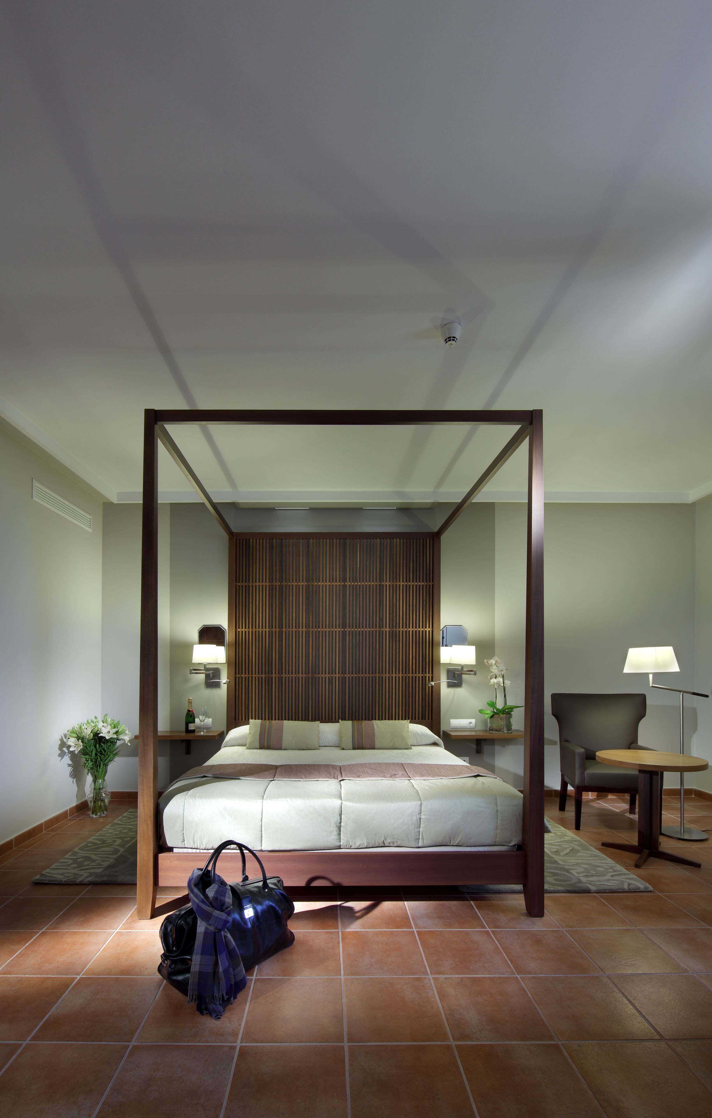 A modern hotel room with a four-poster bed, a wooden headboard, a bedside table with a lamp, a chair, and a small round table.