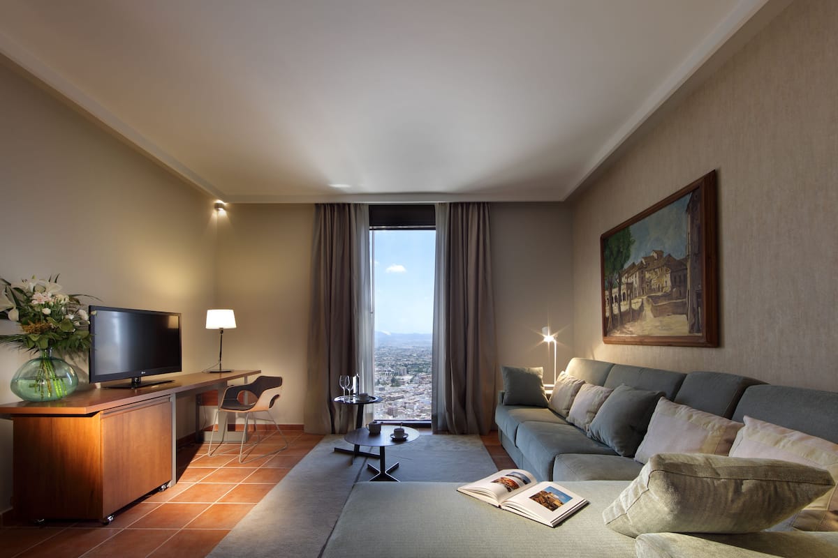 Habitación de hotel moderna con televisor de pantalla plana, escritorio de madera, sofá y ventana grande con vistas a la ciudad.