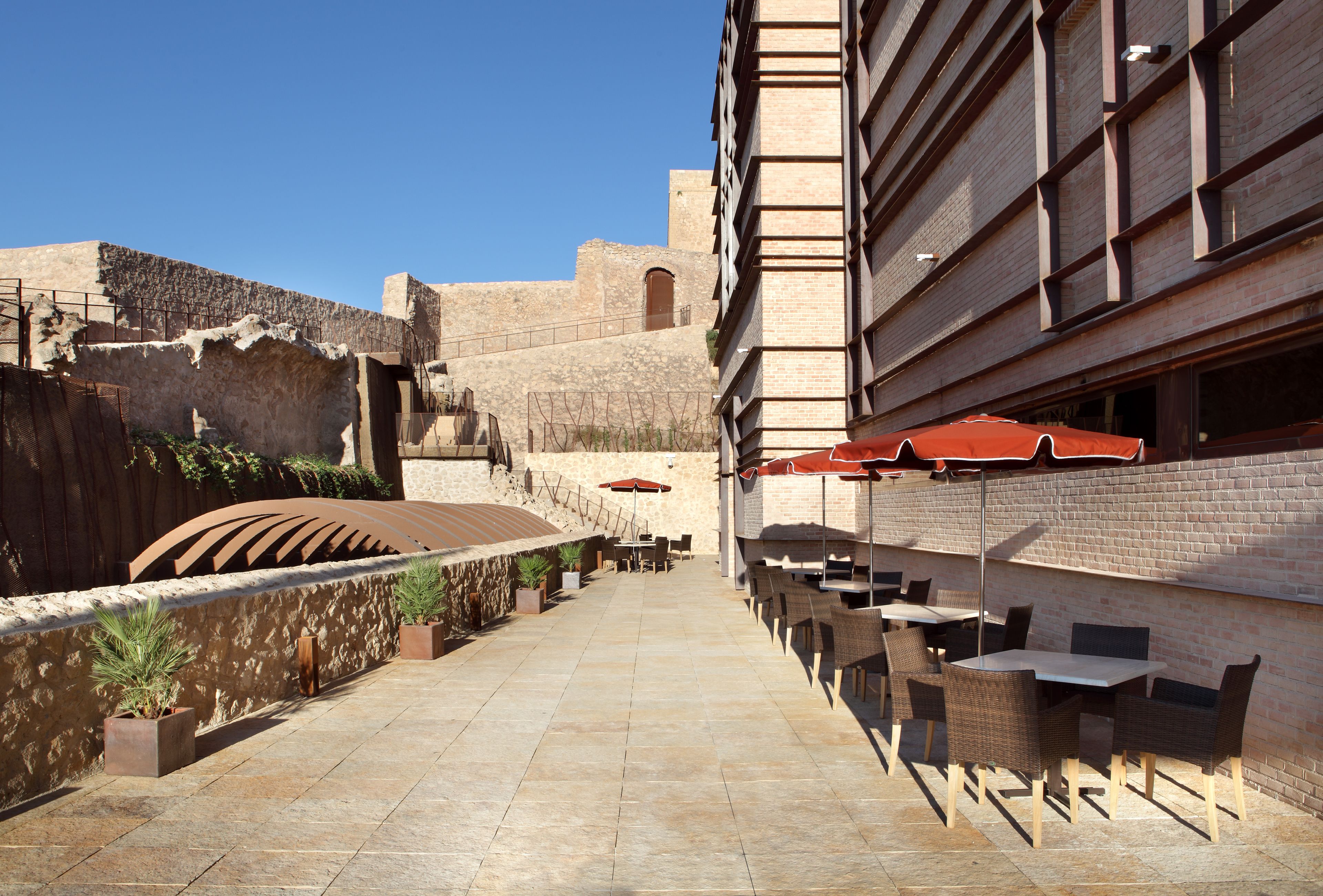 Un patio al aire libre con suelo de piedra, mobiliario de mimbre y sombrillas rojas.