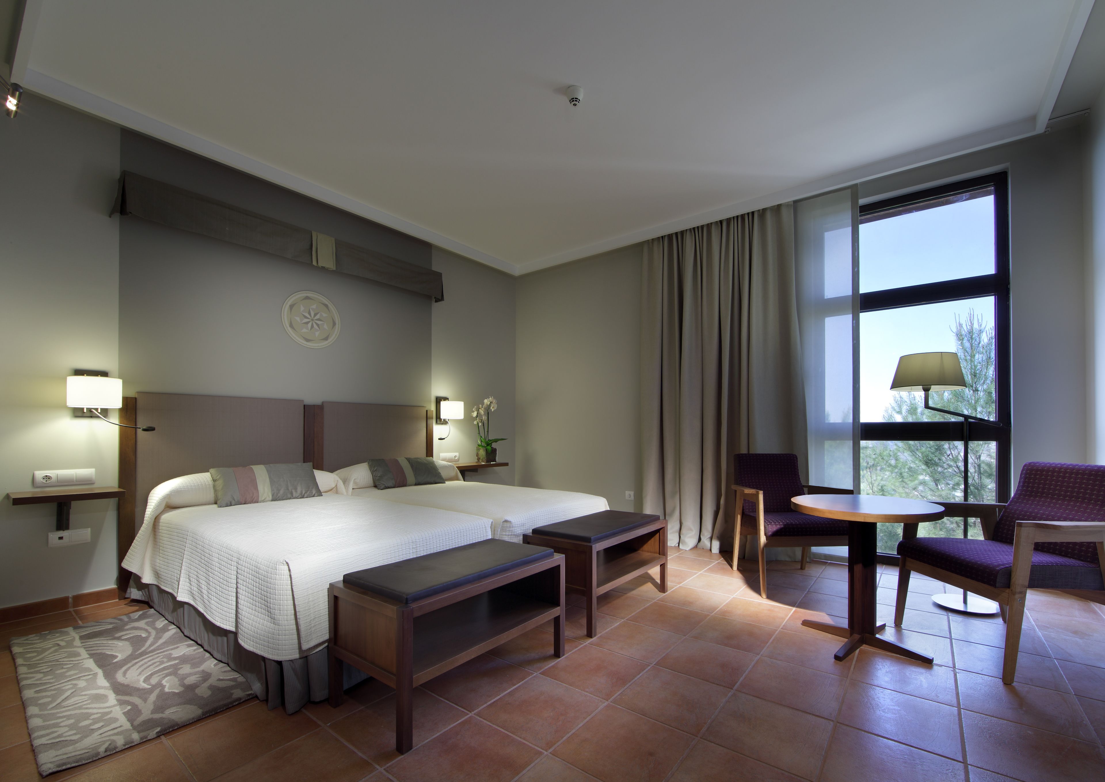 Habitación de hotel con dos camas, un escritorio, una silla, una lámpara y un ventanal con cortinas.