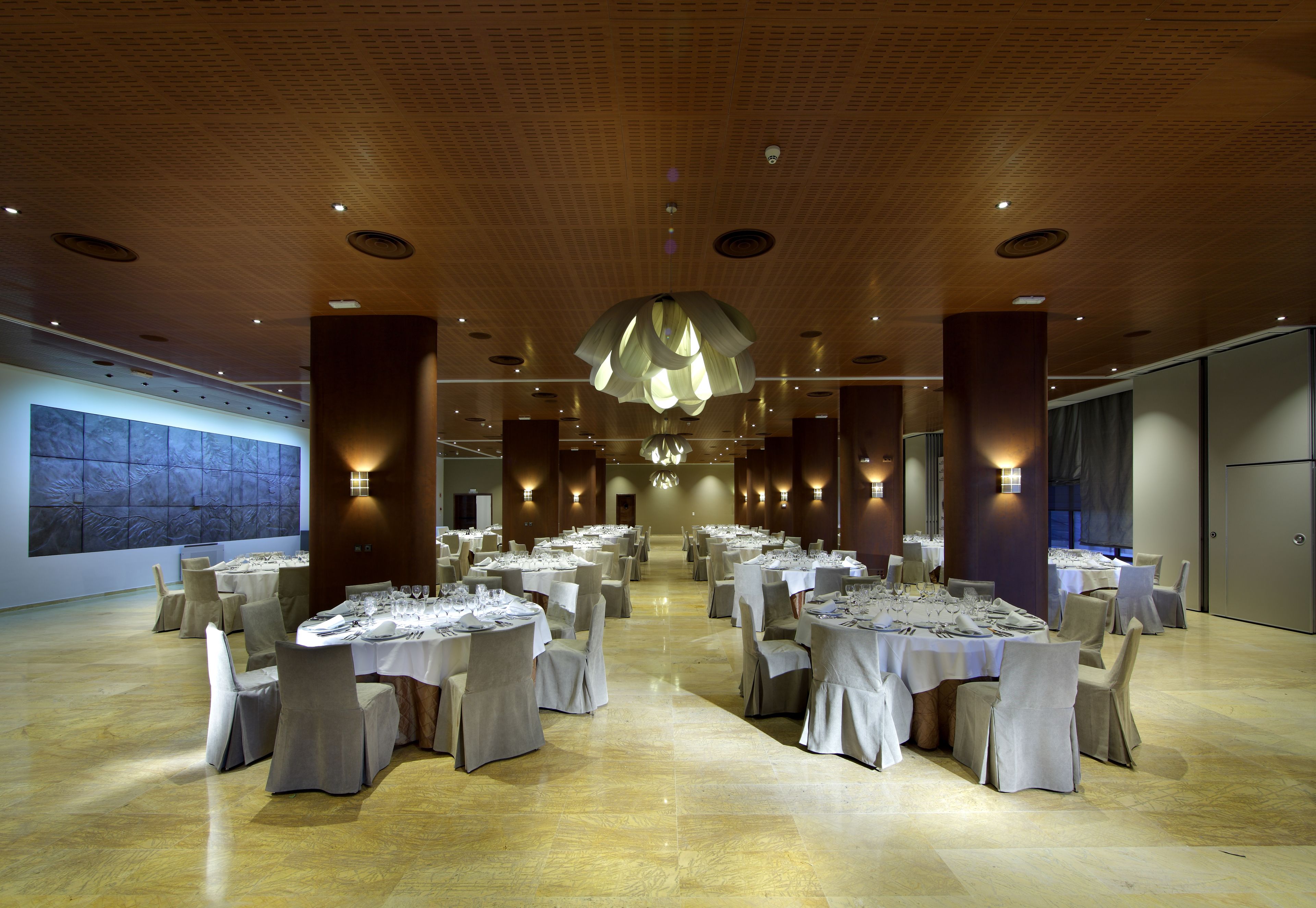 A spacious banquet hall with multiple round tables set for dining, elegant lighting fixtures, and a large abstract mural on the wall.