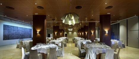 A spacious banquet hall with multiple round tables set for dining, elegant lighting fixtures, and a large abstract mural on the wall.