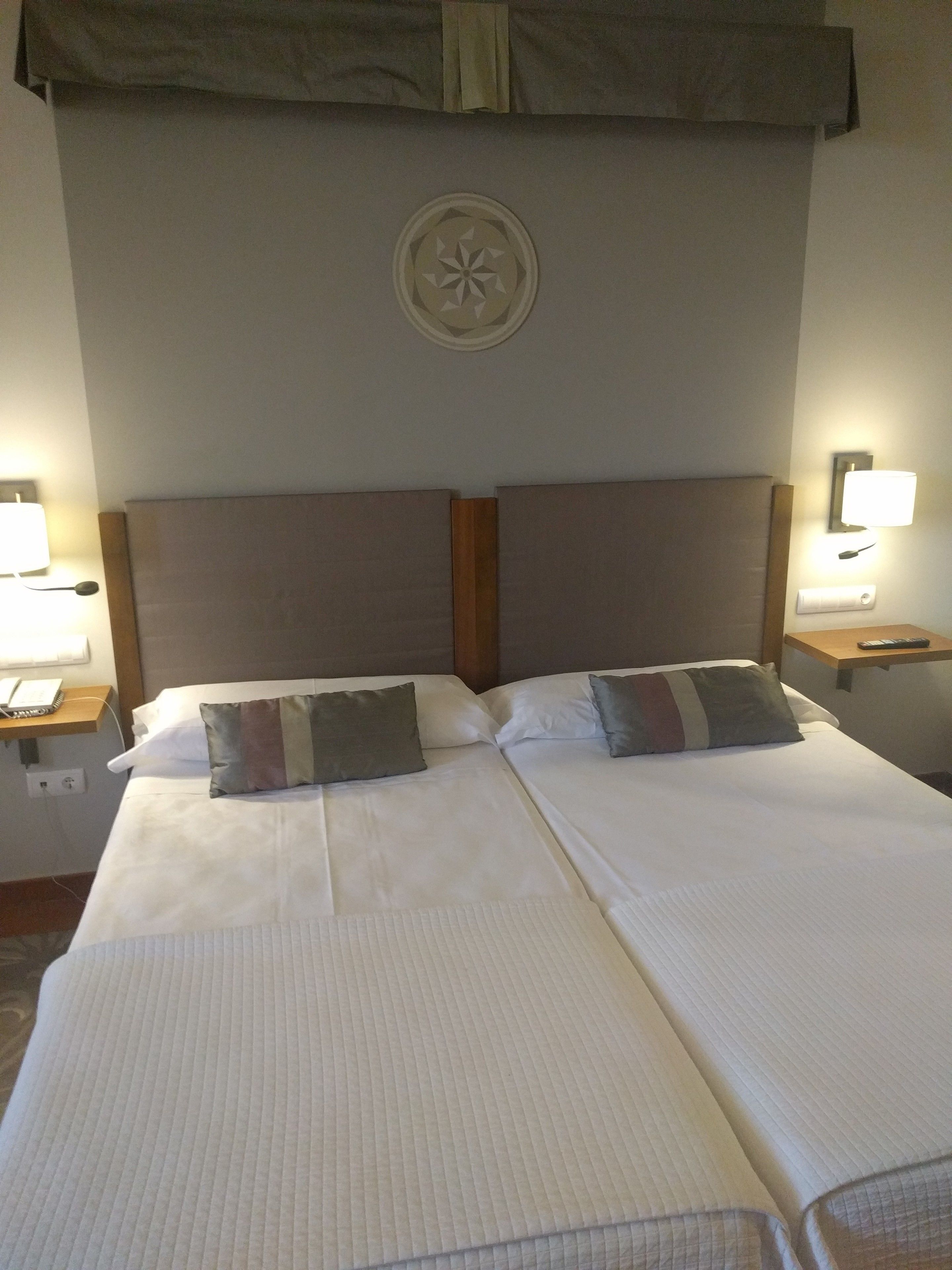 Habitació d'hotel amb dos llits, un capçal amb panell decoratiu, taules de nit amb làmpades i un lluminari fixat a la paret.