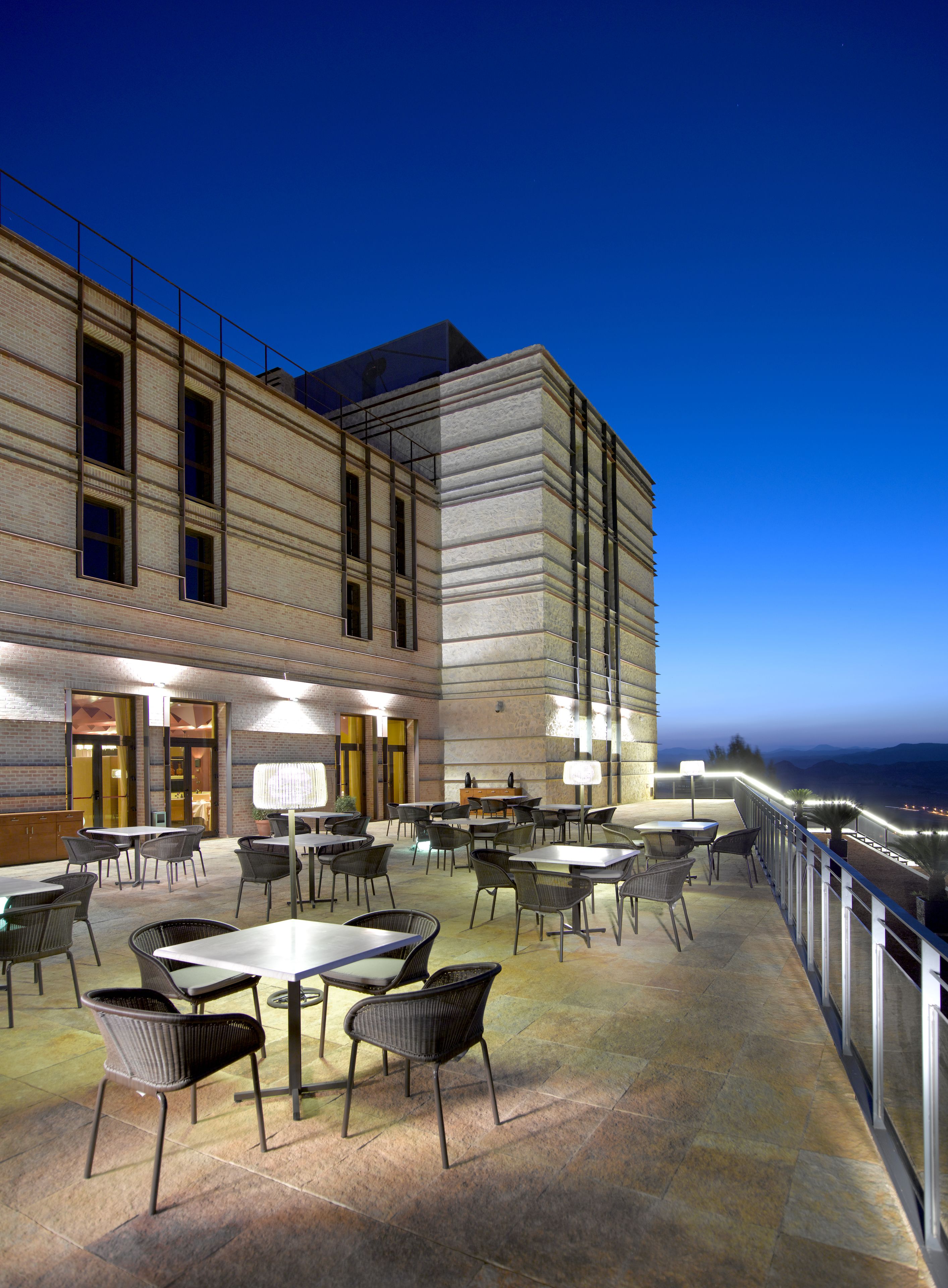 An outdoor patio area with multiple tables and chairs, a brick building with large windows, and a view of the coastline at dusk.