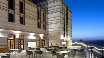 Un patio al aire libre con varias mesas y sillas, un edificio de ladrillo con ventanas amplias y vistas a la costa al anochecer.