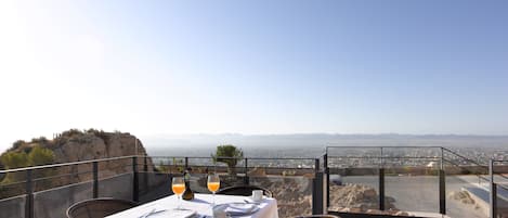 A dining table set with white tablecloth, wicker chairs, and a glass railing overlooking a cityscape and mountains.
