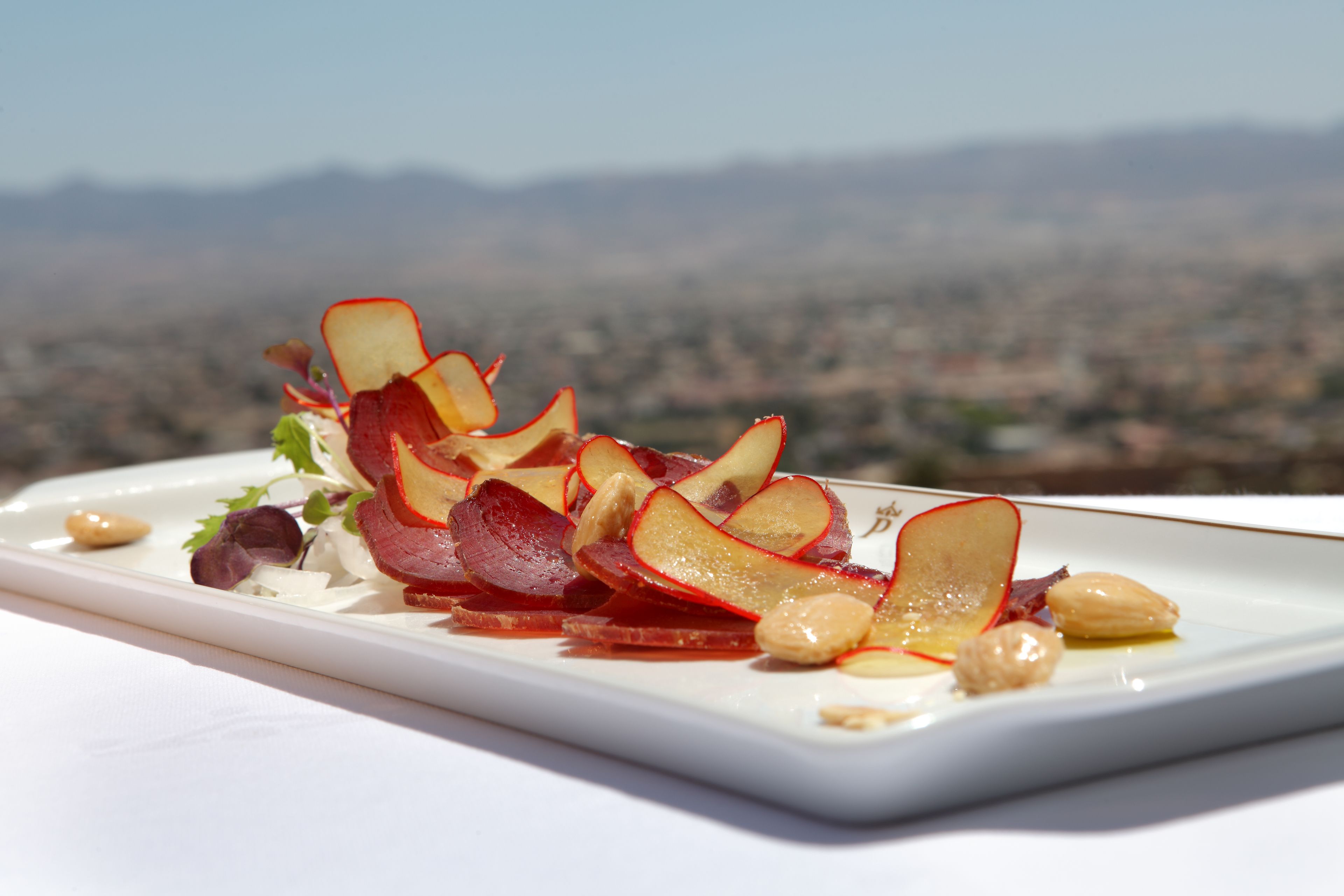 A white rectangular plate with a food arrangement featuring apple slices and nuts, set against a blurred mountain backdrop.