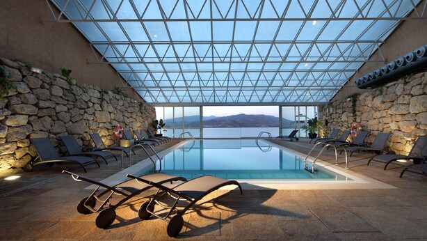 An indoor swimming pool with a glass ceiling, stone walls, and lounge chairs. - Parador de Lorca (Lorca)