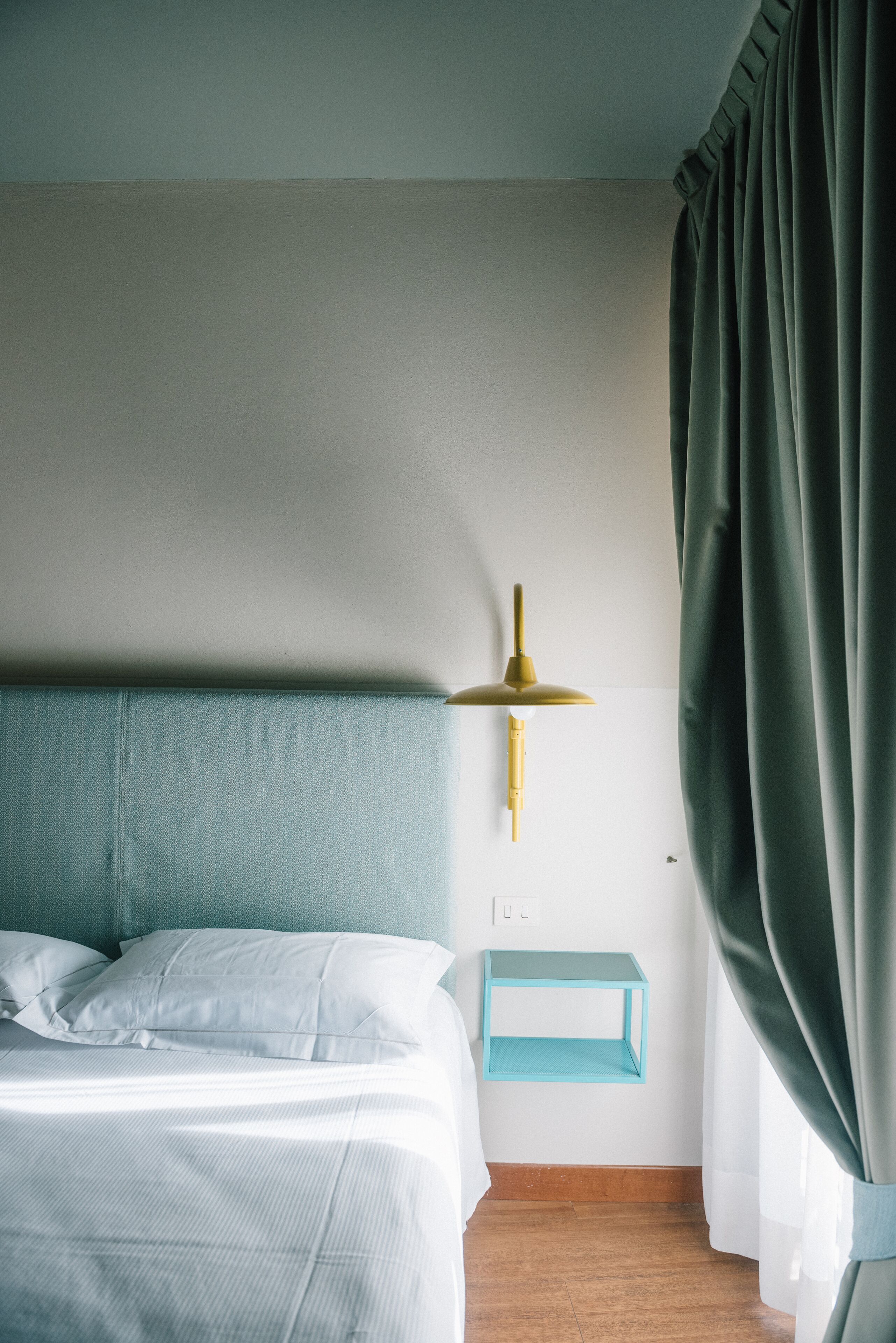 A neatly made bed with white linens, a teal headboard, a gold lamp on a white wall, and a teal side table.