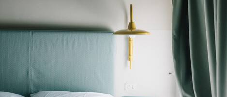 A neatly made bed with white linens, a teal headboard, a gold lamp on a white wall, and a teal side table.