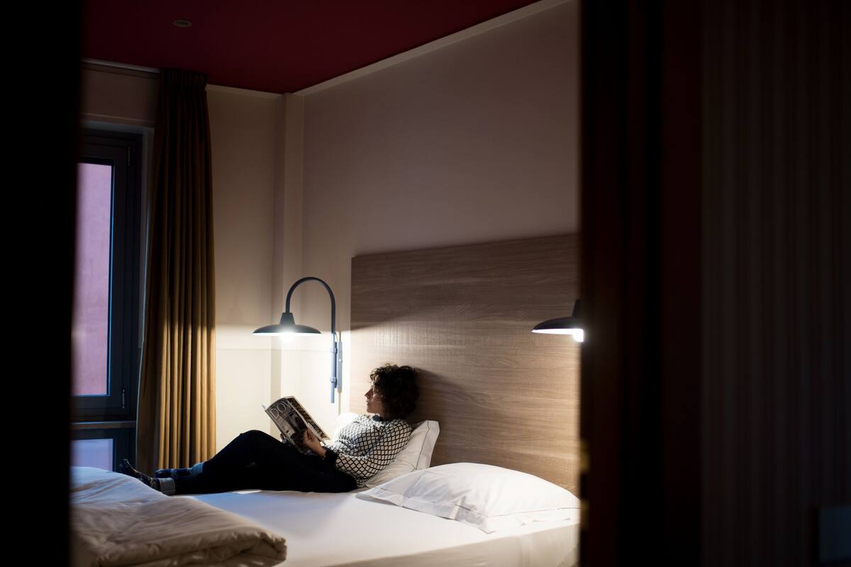 A hotel room with a bed, a reading lamp, and a window with curtains.