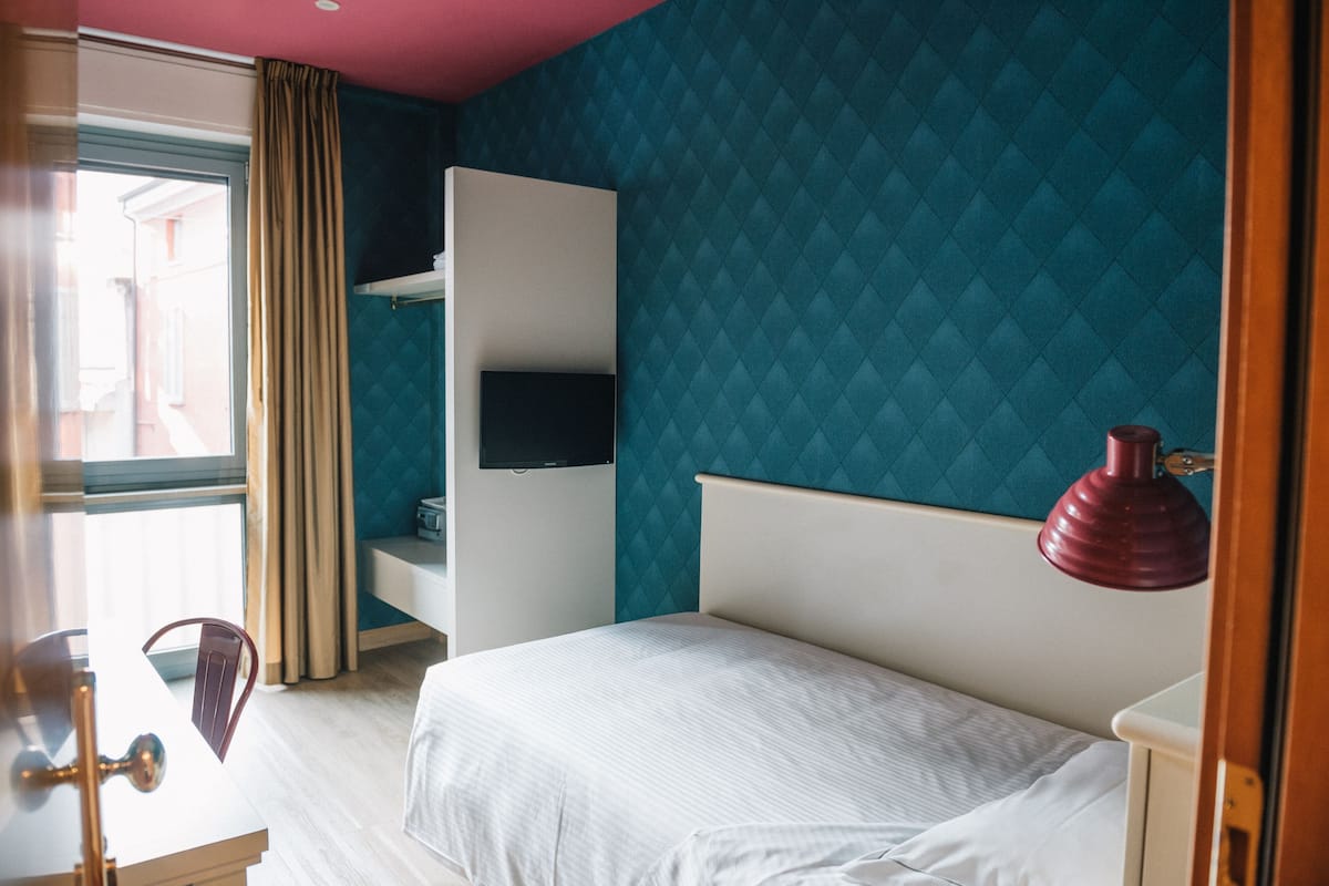 A hotel room with a bed, a TV mounted on the wall, a desk with a chair, and a window with curtains.