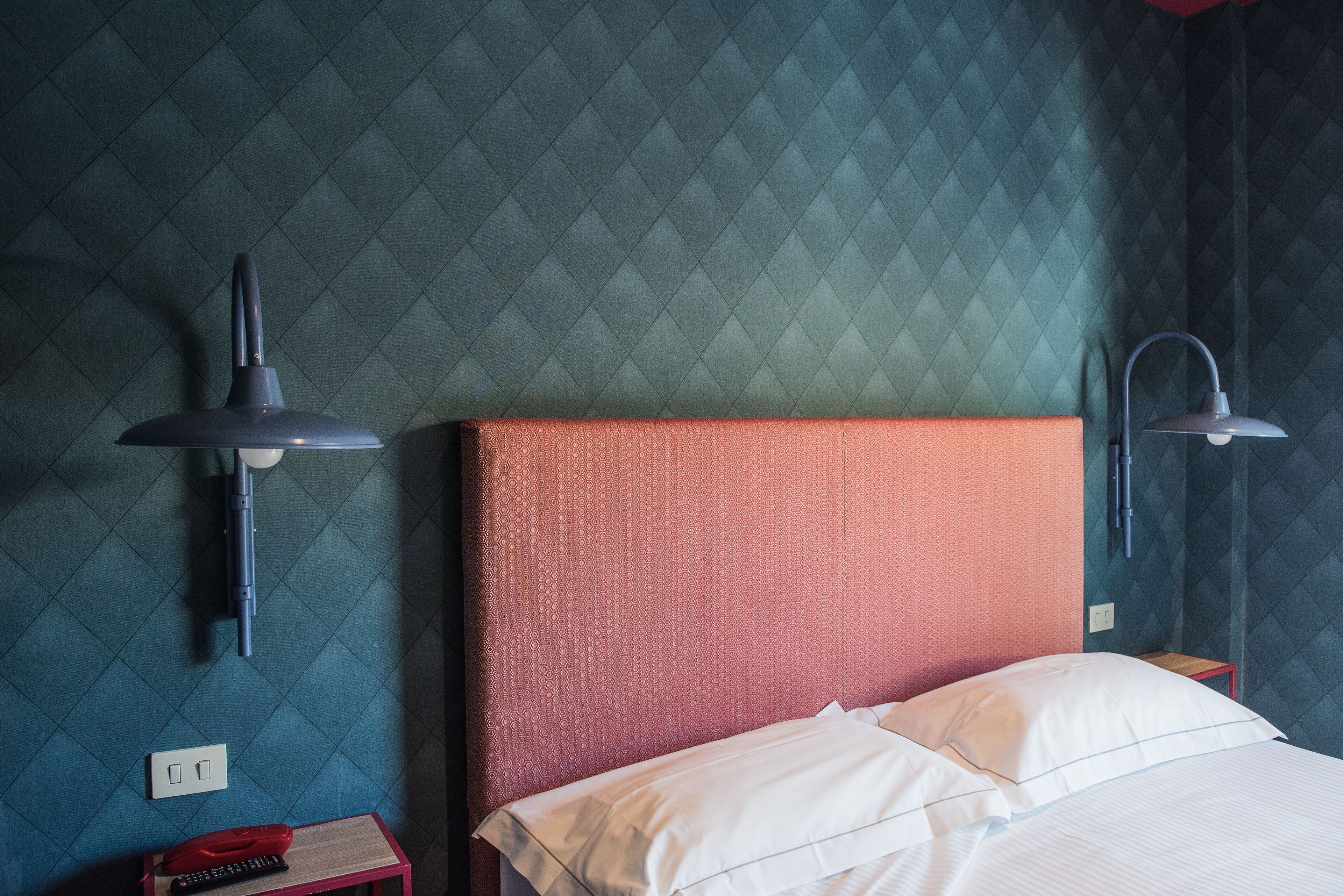 Quarto de hotel com uma cama, duas luminárias na parede, uma mesa de cabeceira e um telefone vermelho.