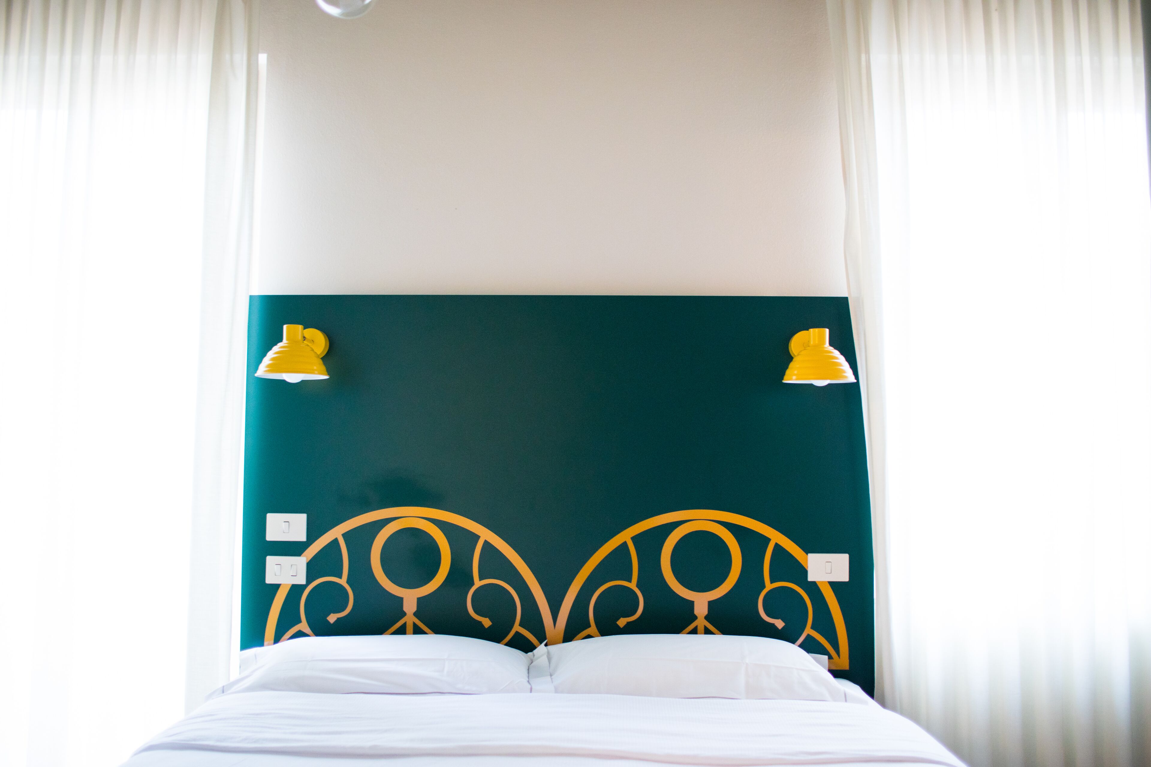Ein Bett mit weißer Bettwäsche, einem türkisblauen Kopfteil und zwei an der Wand befestigten gelben Lampen.
