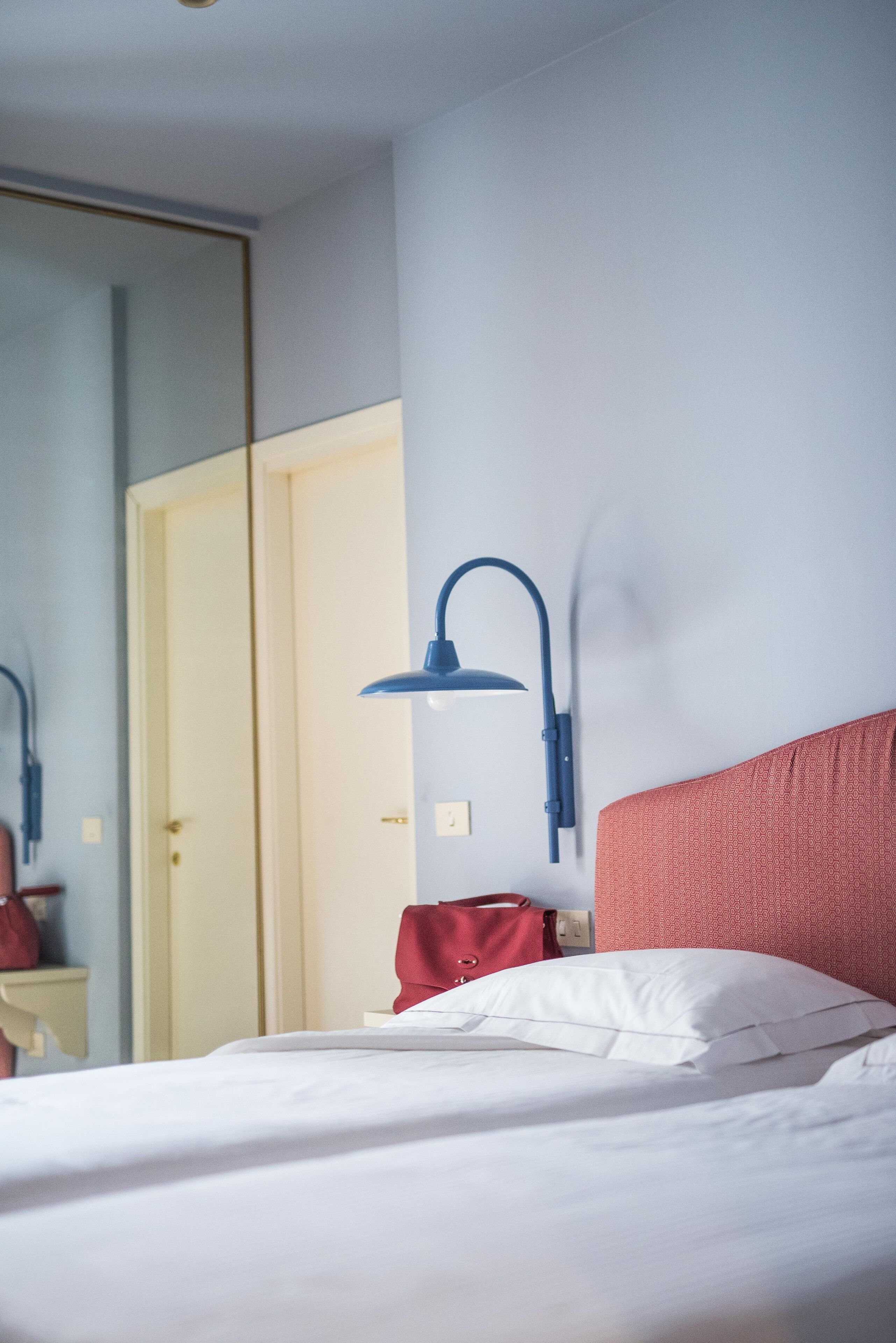 Una habitación de hotel con una cama, cabecera roja, lámpara de pared azul y una puerta al fondo.