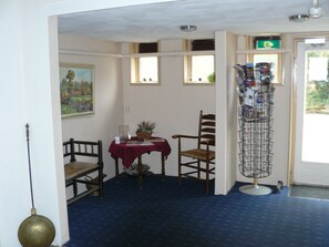 A room with a blue carpet, a table with a red tablecloth, two wooden chairs, a potted plant, a wall-mounted shelf with magazines, and a painting.