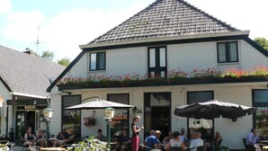 An outdoor seating area with tables and chairs, a bicycle, and a building with a tiled roof and flower boxes. - De Koningsherberg (Anloo)